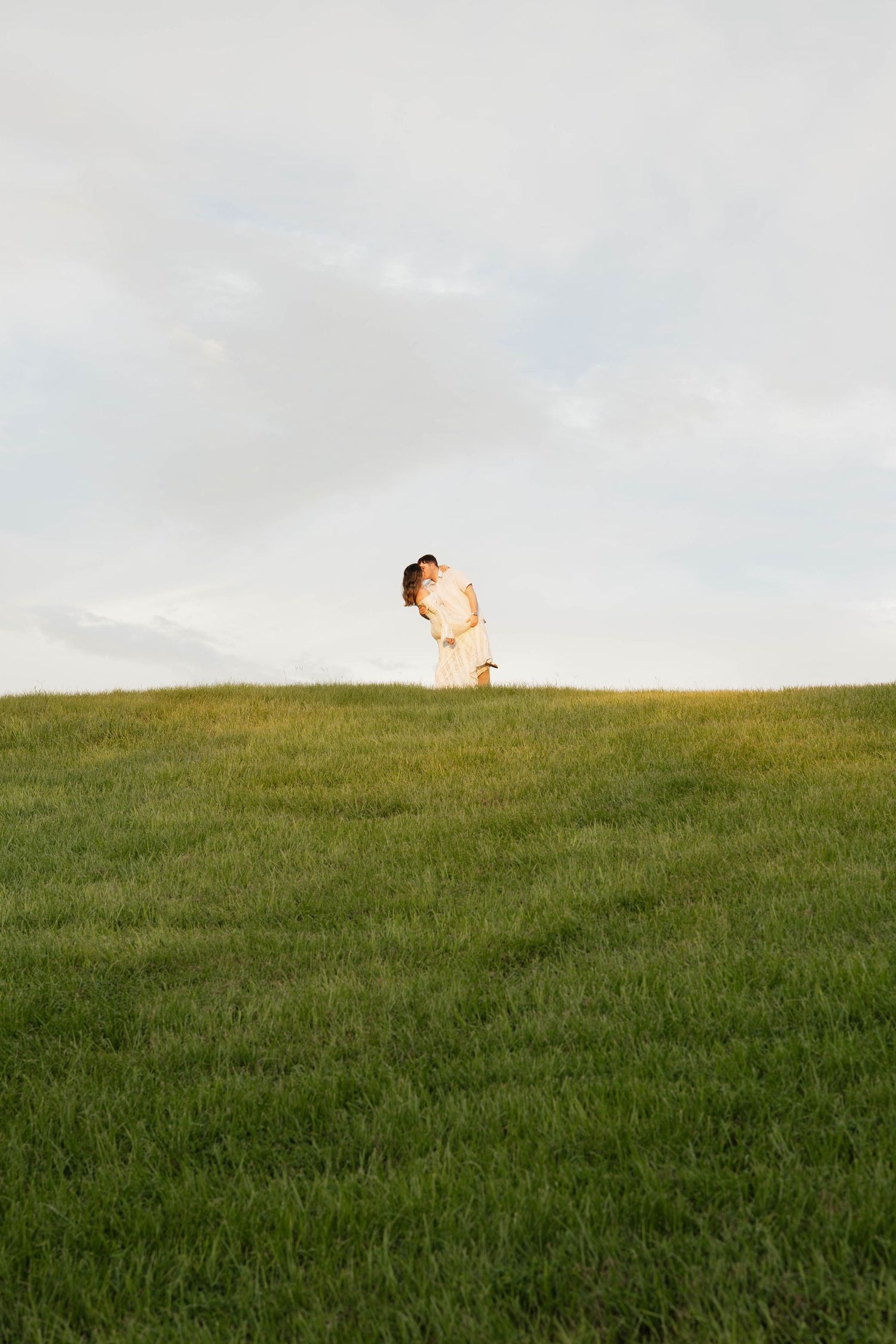 A couple embraces on a lush green hillside under a soft cloudy sky during golden hour.