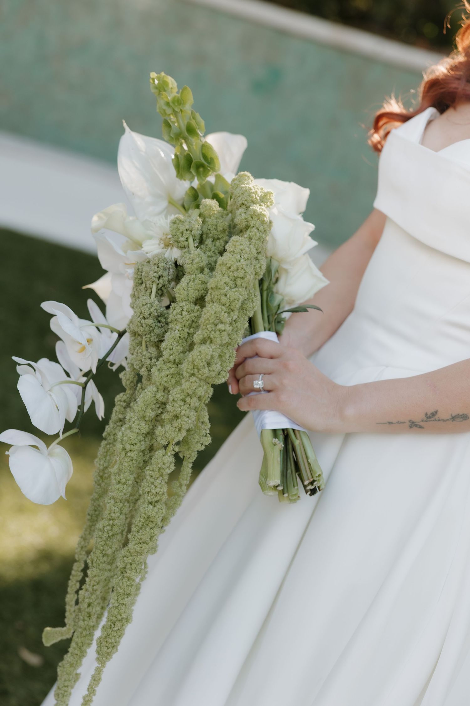 Bride in white gown holding an elegant cascading bouquet of white flowers and green amaranthus near a pool.