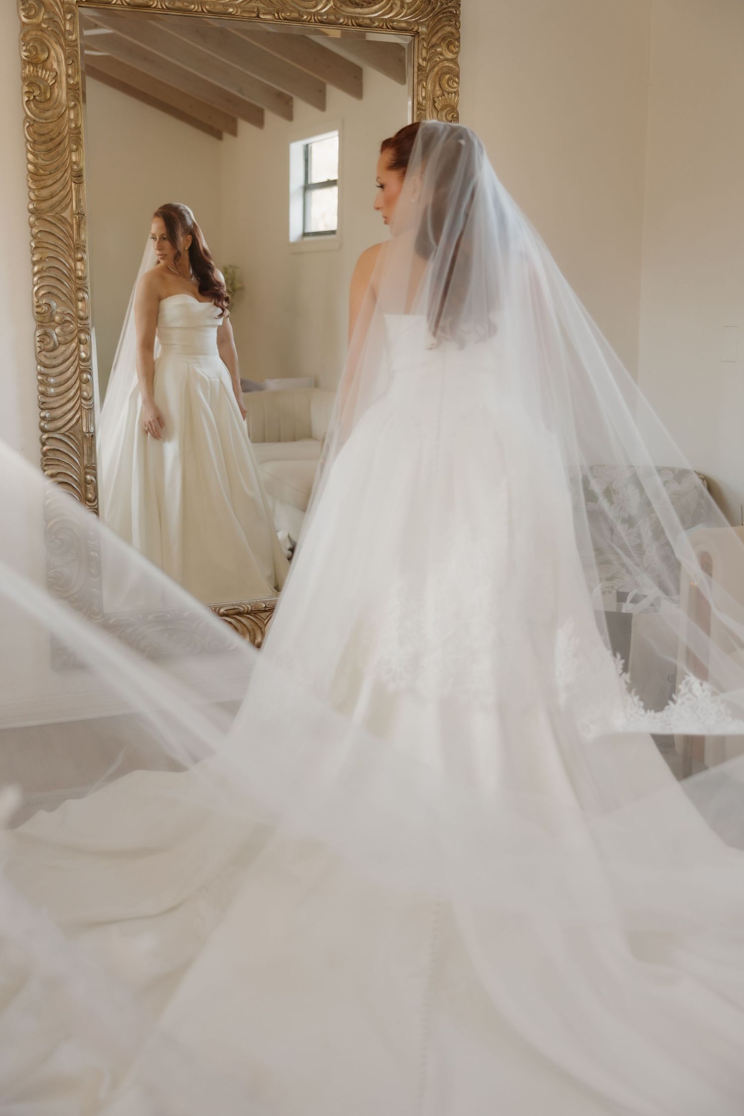 A bride in an elegant white wedding gown and flowing veil looks at her reflection in an ornate gold-framed mirror.