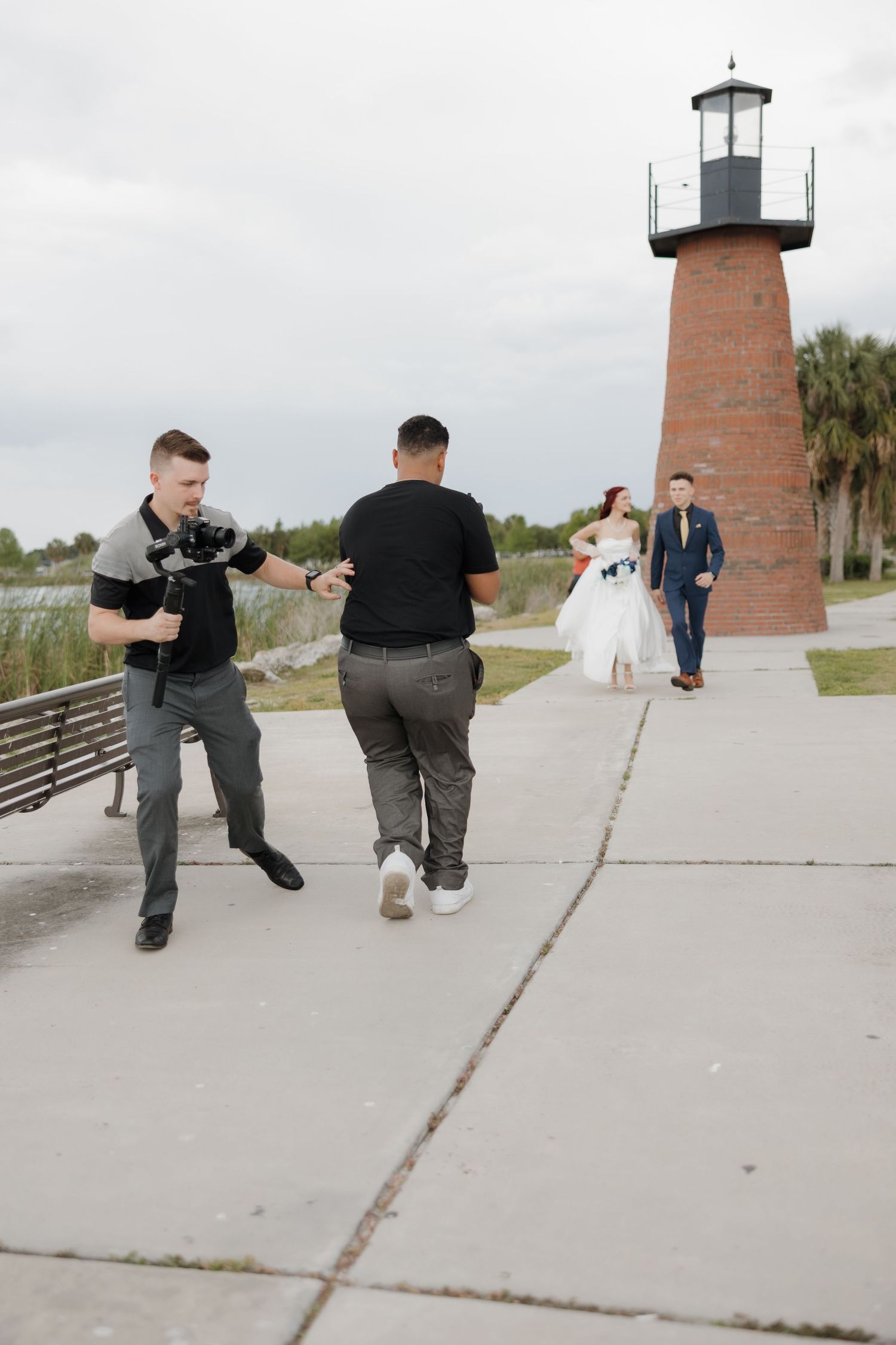 Wedding photographers capture bride and groom walking toward them near a brick lighthouse on a cloudy day.