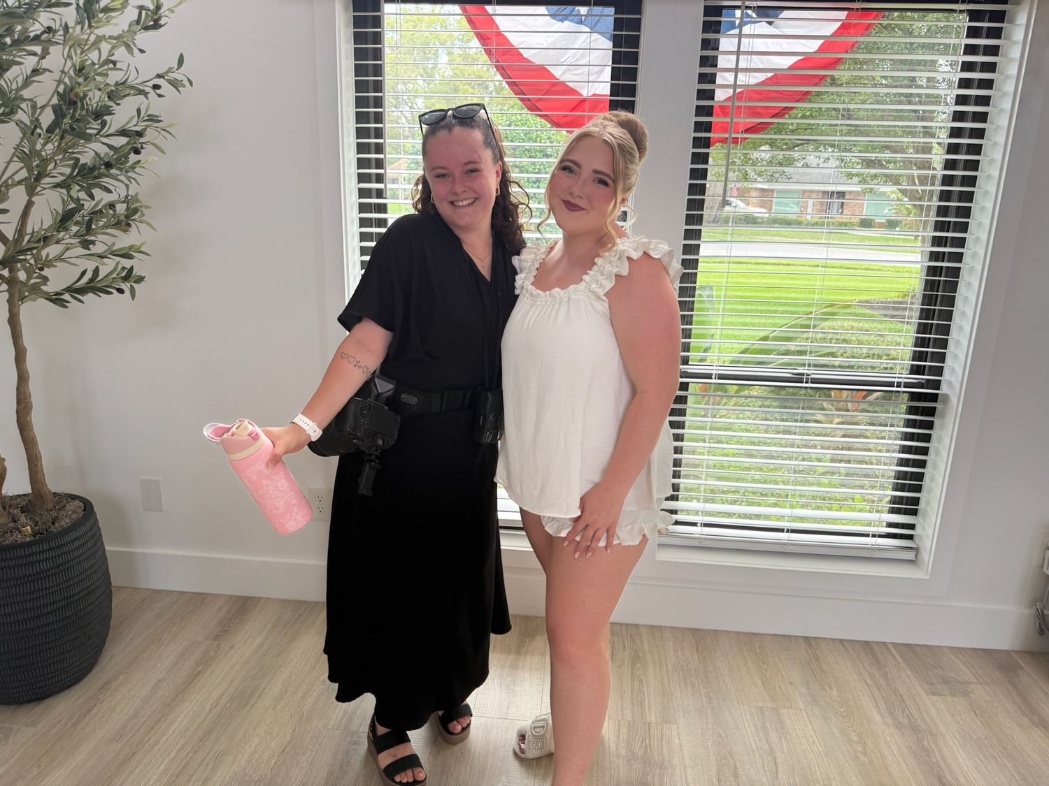 Two women smiling together indoors, one in black dress and one in white outfit, posing near a window.