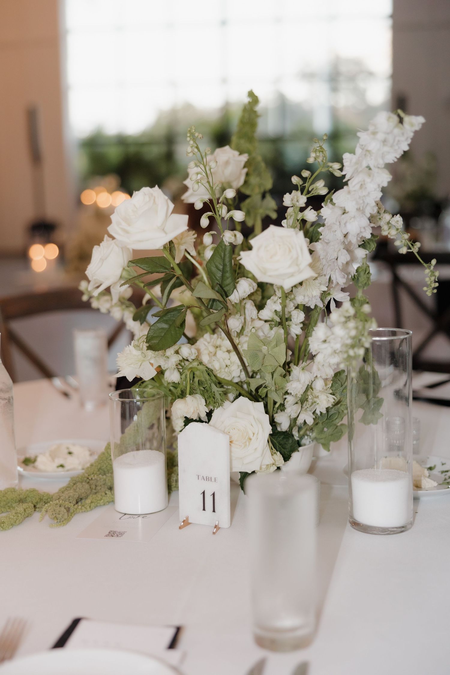 Elegant white floral centerpieces with greenery in frosted glass vases on white tablecloths at a wedding reception.