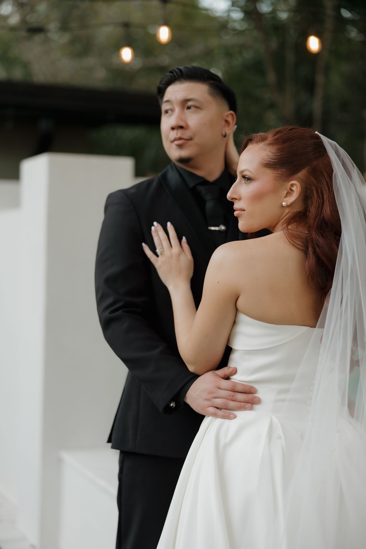 A bride in a white strapless gown and groom in black suit embrace during an outdoor wedding ceremony.