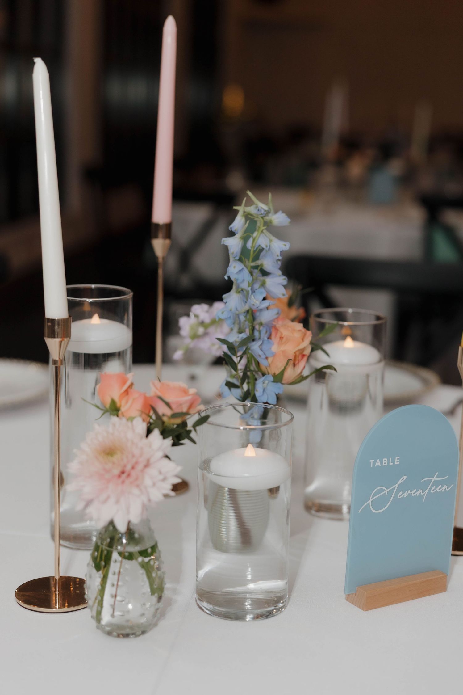Elegant wedding table setting with white candles, soft pink flowers, and light blue place cards on white linens.