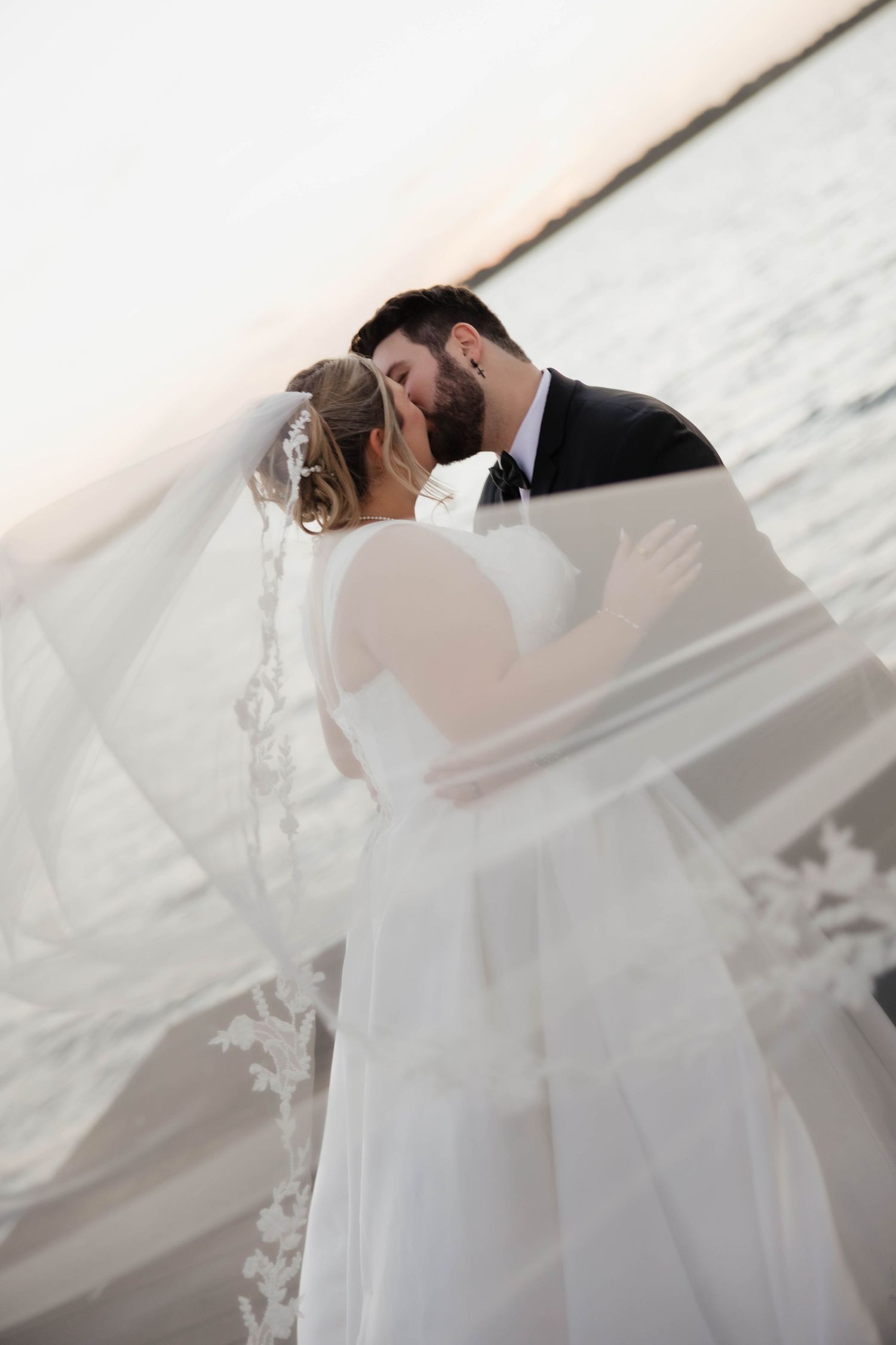 Romantic wedding couple embraces under a flowing white veil during sunset at the beach.