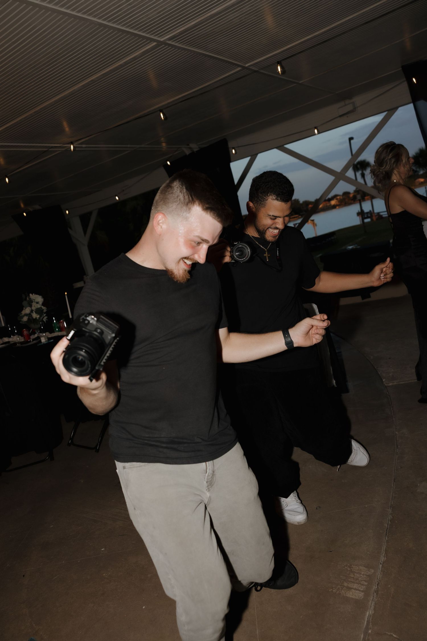 Two men in black shirts dancing at an outdoor evening event, one holding a camera.