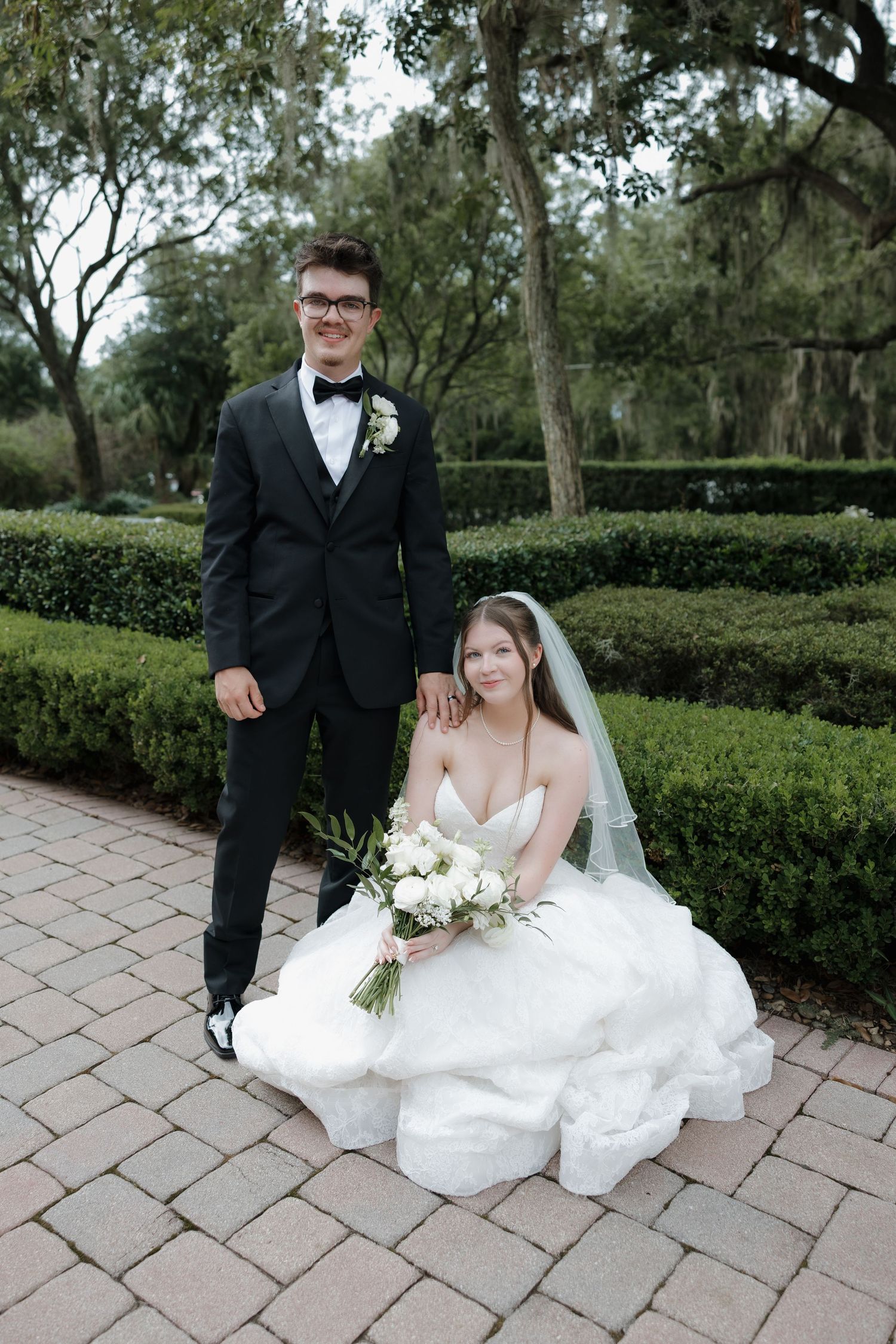 A bride in a white gown and veil sits holding a bouquet while her groom in a black tuxedo stands beside her outdoors.