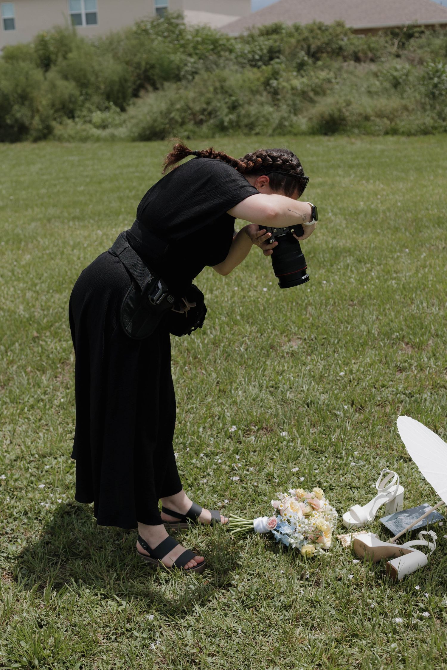 Photographer in black dress bending down to capture bouquet and shoes on green grass outdoors.