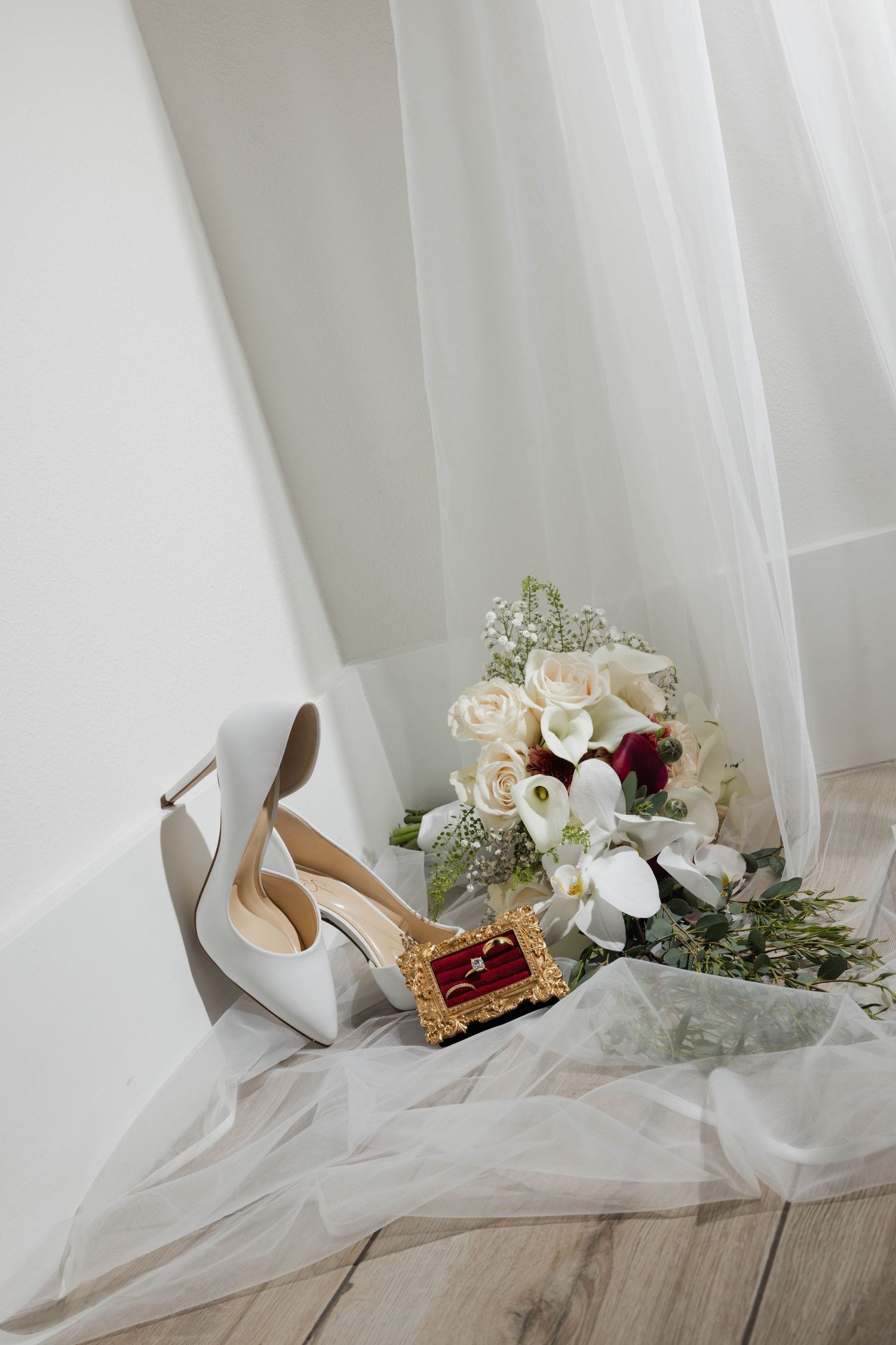 White bridal heels, gold ring box, and elegant bouquet of roses arranged on sheer veil near window.