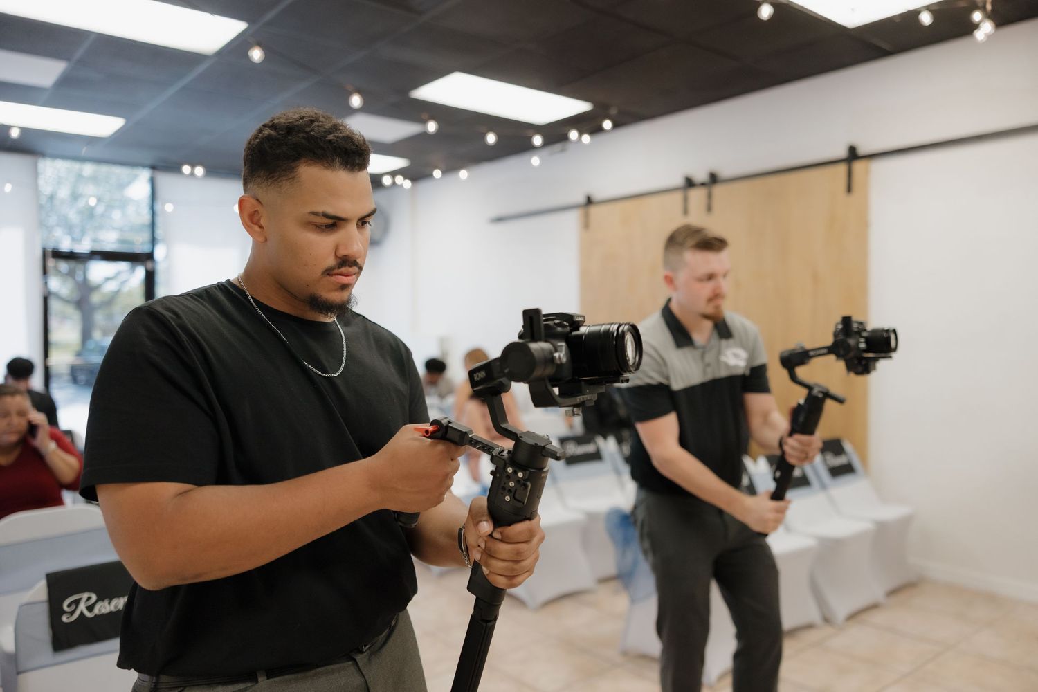 Two videographers holding camera gimbals practice filming techniques in a bright modern studio space.