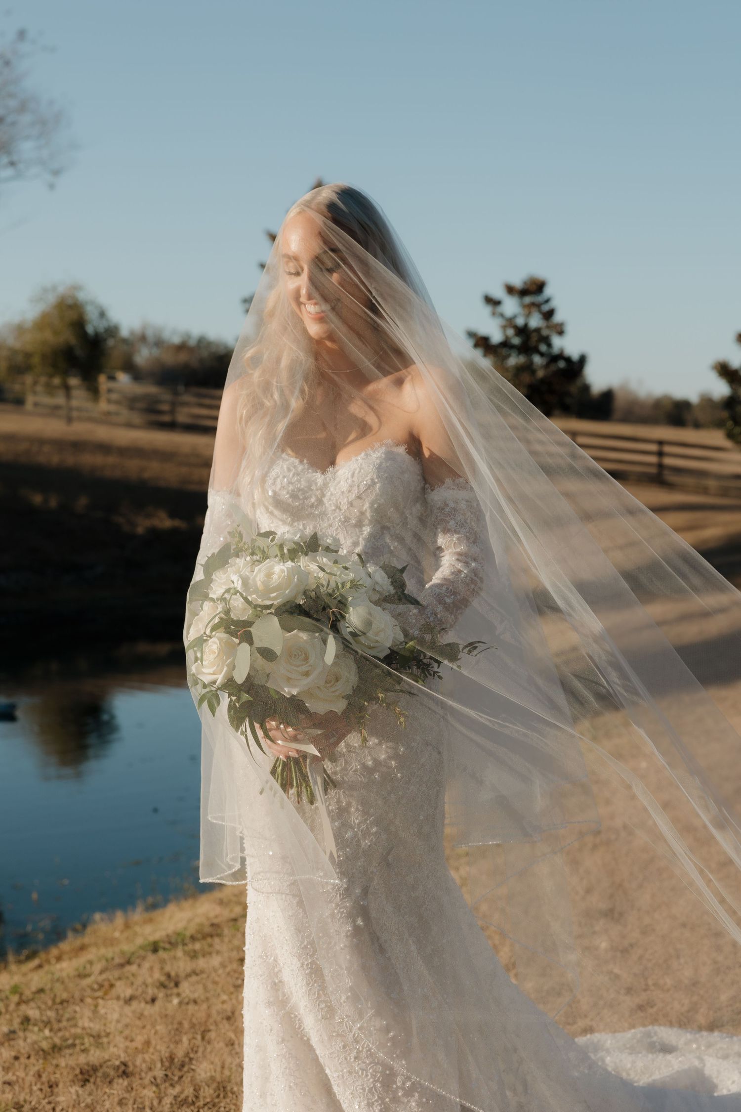 A bride in a white lace gown holds a bouquet of white flowers as her veil flows in the breeze by a lake at sunset.