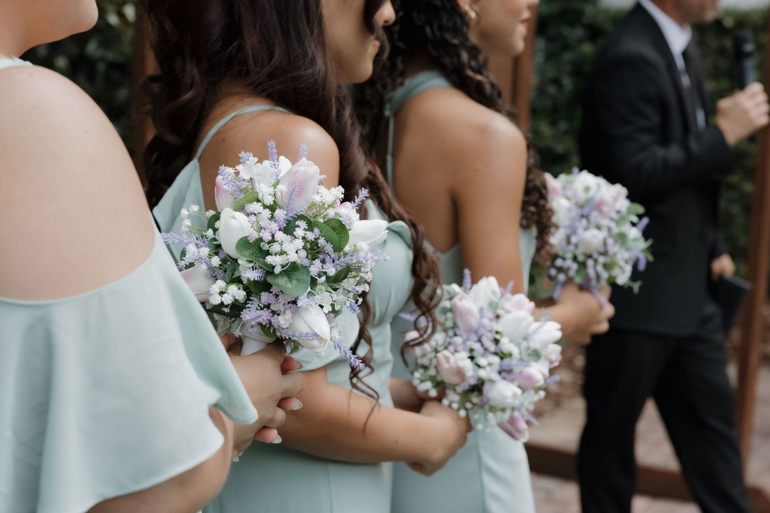 Bridesmaids in mint green dresses holding small bouquets of purple and white flowers at a wedding ceremony.