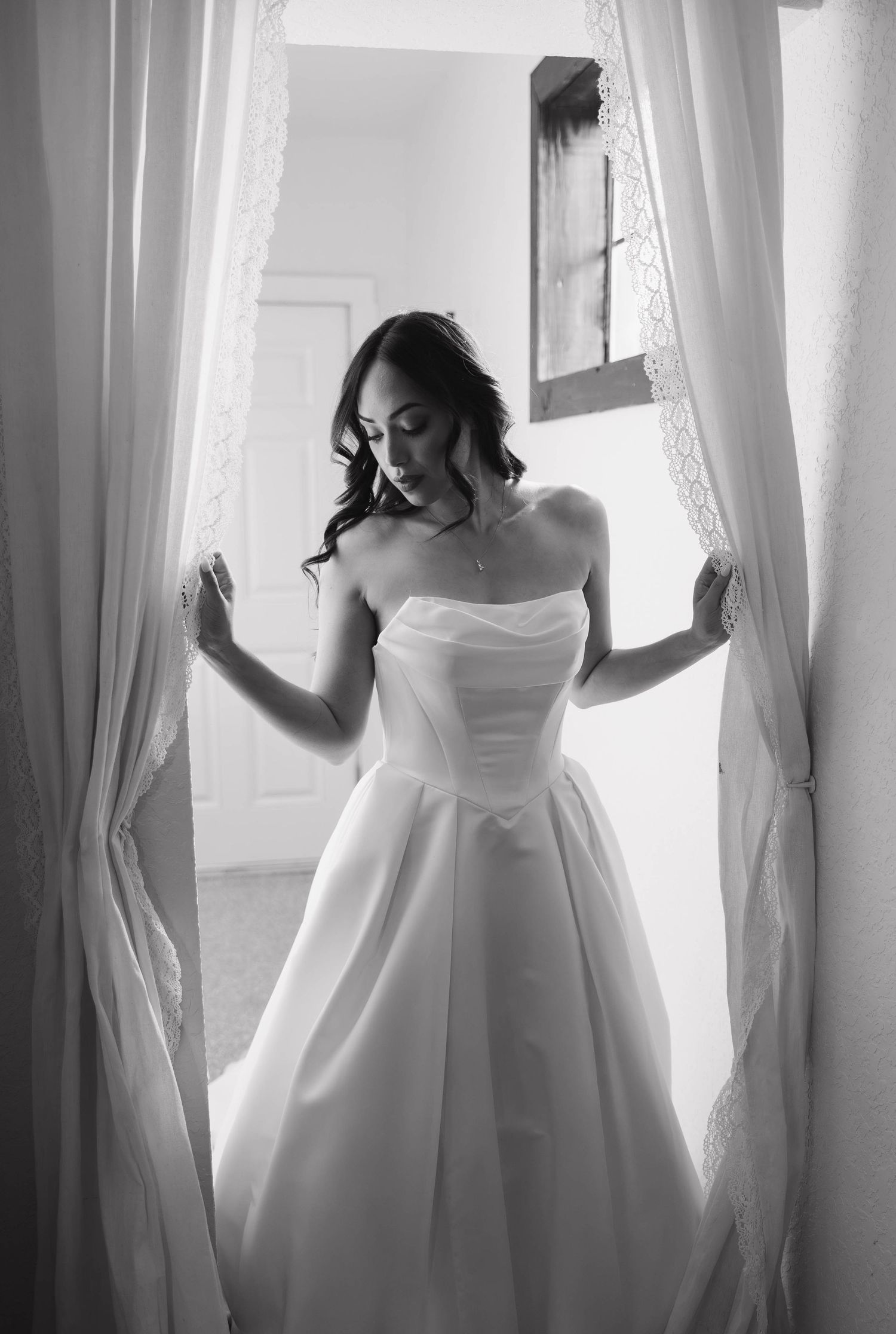 Black and white photo of a bride in a strapless ball gown standing between sheer curtains in soft window light.