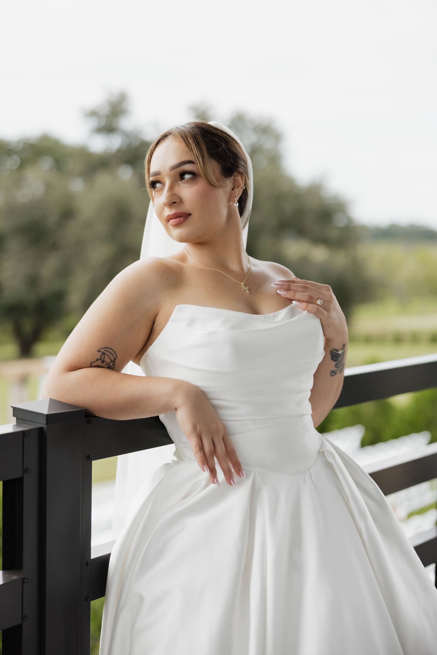 Bride in strapless white ball gown with veil and tattoos poses on outdoor railing at elegant wedding venue.