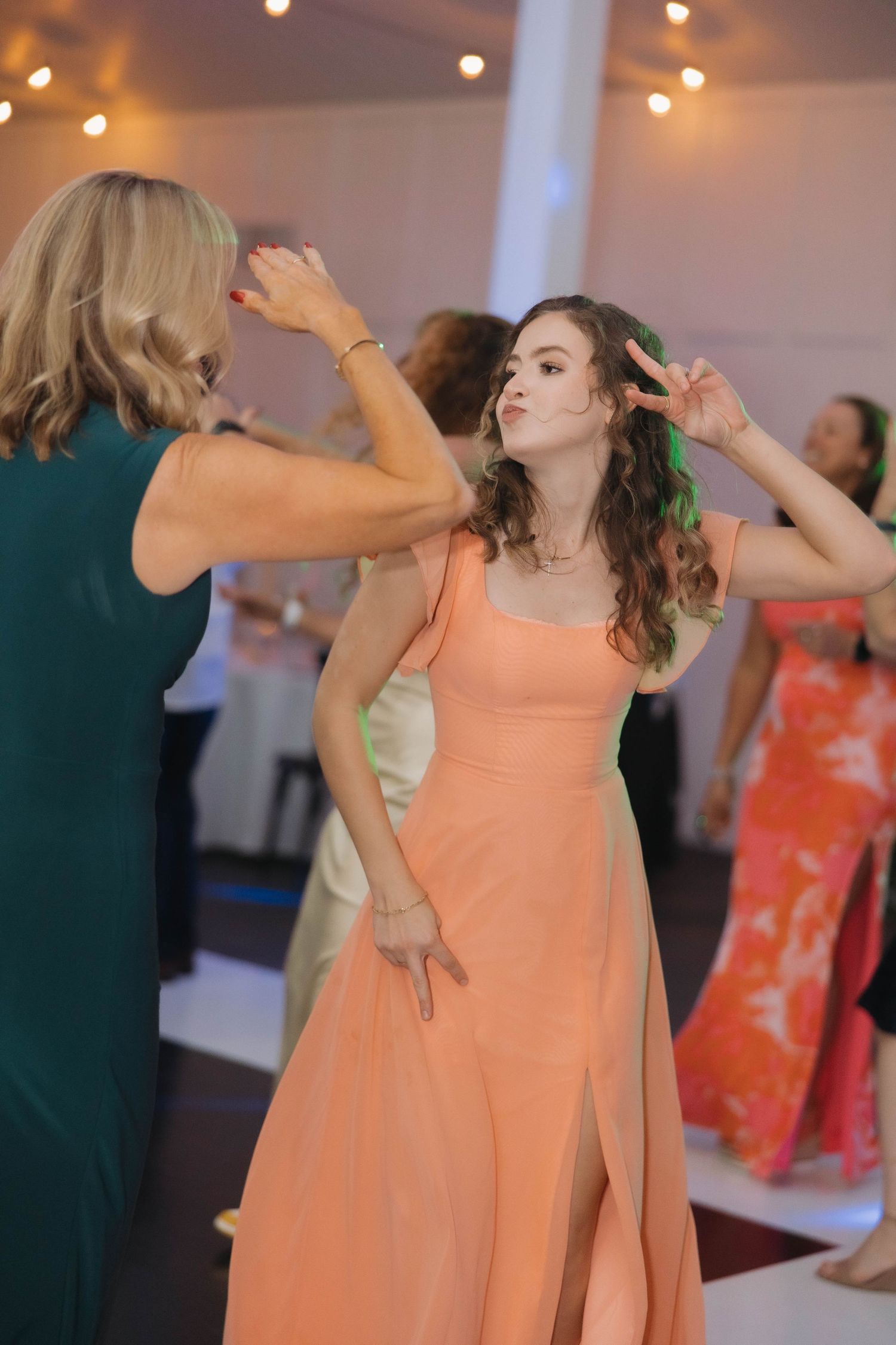 People dancing and having fun at an indoor celebration with string lights, featuring a woman in a flowing peach-colored dress.