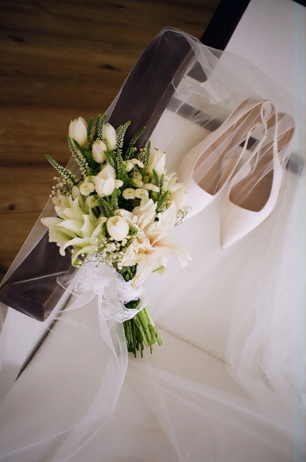 Elegant bridal bouquet of white tulips and lilies beside satin heels and tulle veil on a wooden floor.