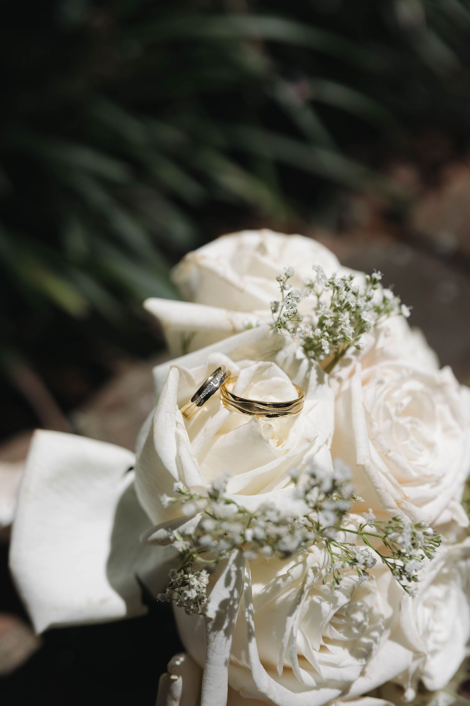 Gold wedding rings resting on a bridal bouquet of white roses and baby's breath, with green foliage background.