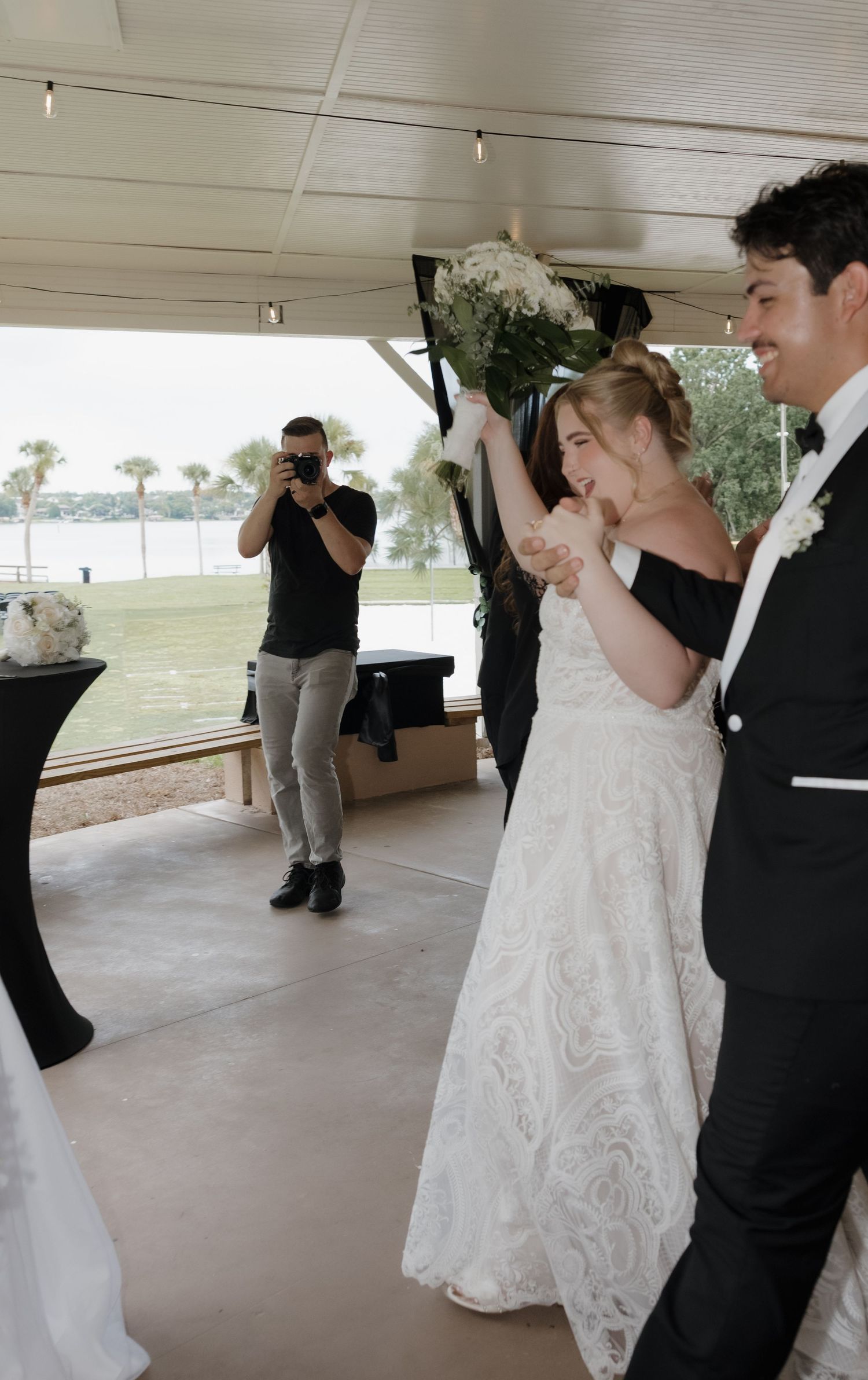 Joyful bride in white lace gown raises bouquet with groom in tuxedo as photographer captures wedding ceremony moment.