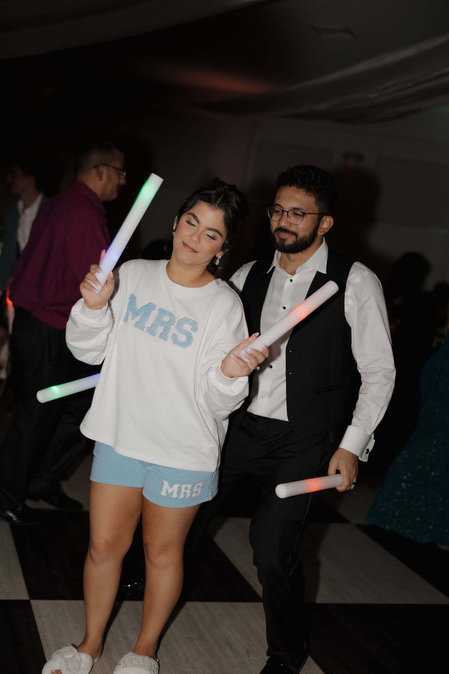 Two people at a party hold glowing light sticks while dancing, with one wearing a white Mrs sweater and blue shorts.