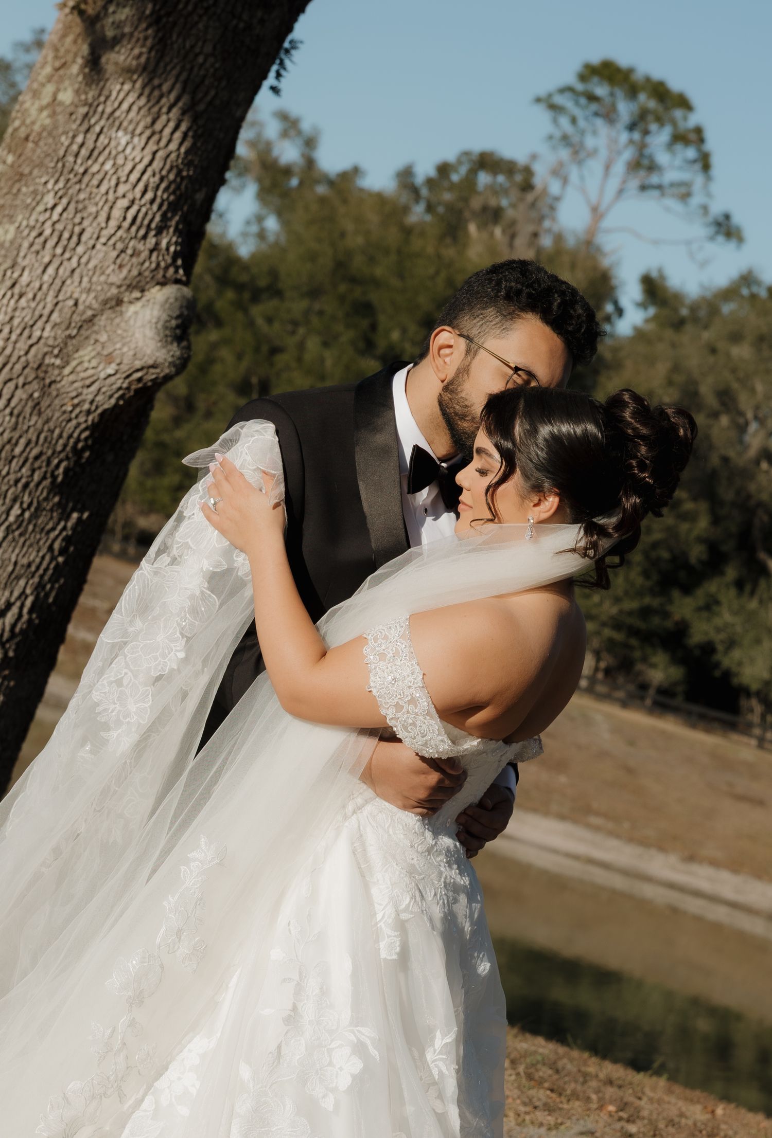 A romantic wedding couple embraces under a tree by a lake during a golden sunset moment.