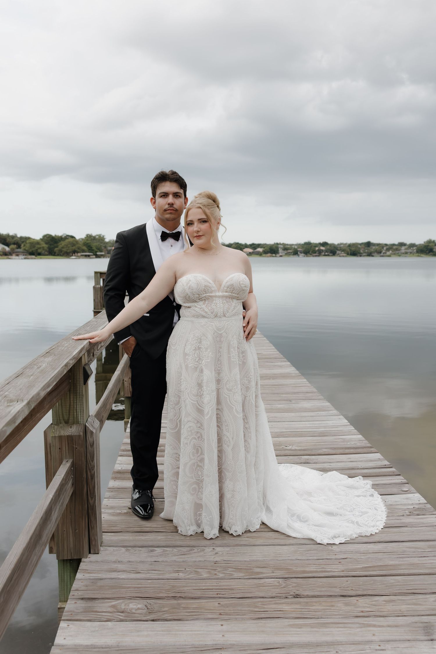 Bride in lace gown and groom in tuxedo pose on a wooden dock overlooking a calm lake on a cloudy day.