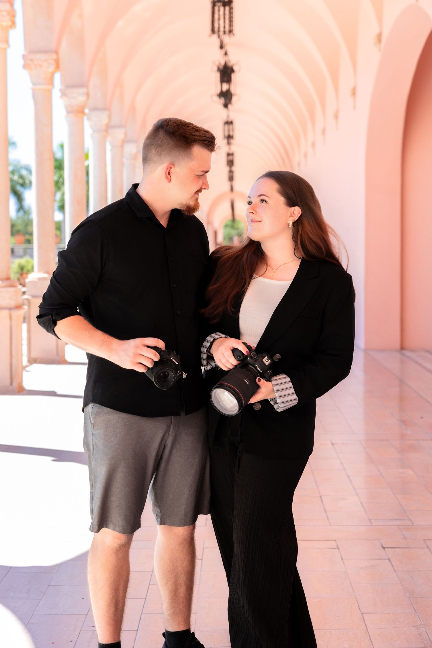 A couple holding cameras smiles at each other in a sunlit pink archway corridor.