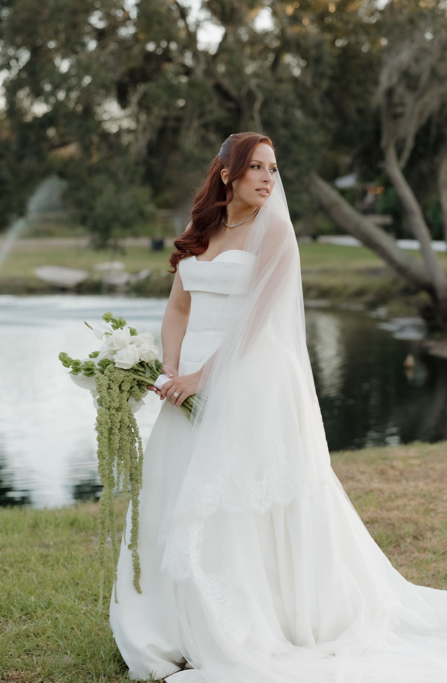 A bride in a flowing white wedding dress and veil stands beside a lake holding a cascading bouquet of white flowers.