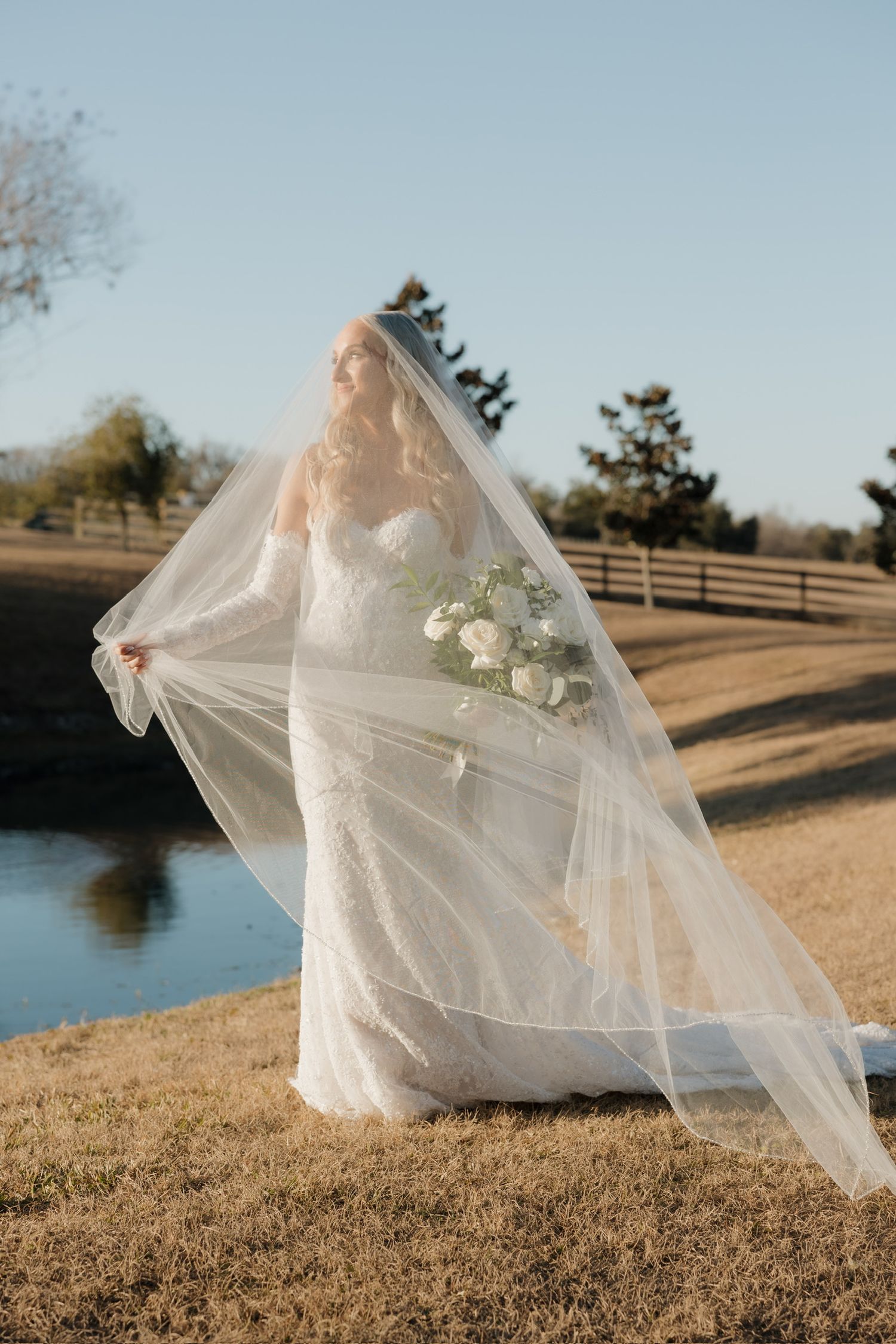 Bride in lace gown holding white bouquet with flowing veil outdoors near pond on sunny day.