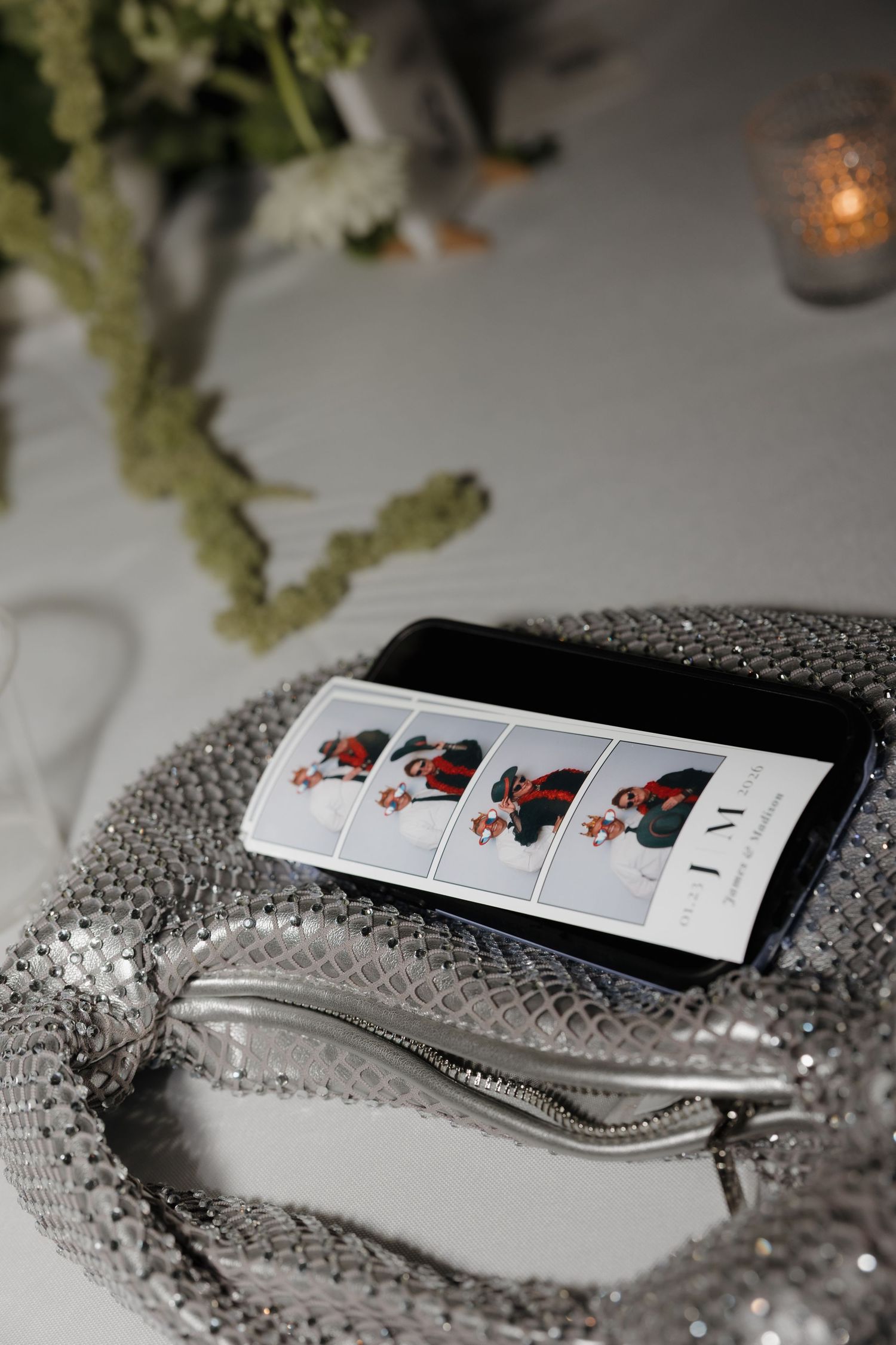 A silver sequined handbag with a phone displaying photo booth style pictures next to a candlelit table setting.