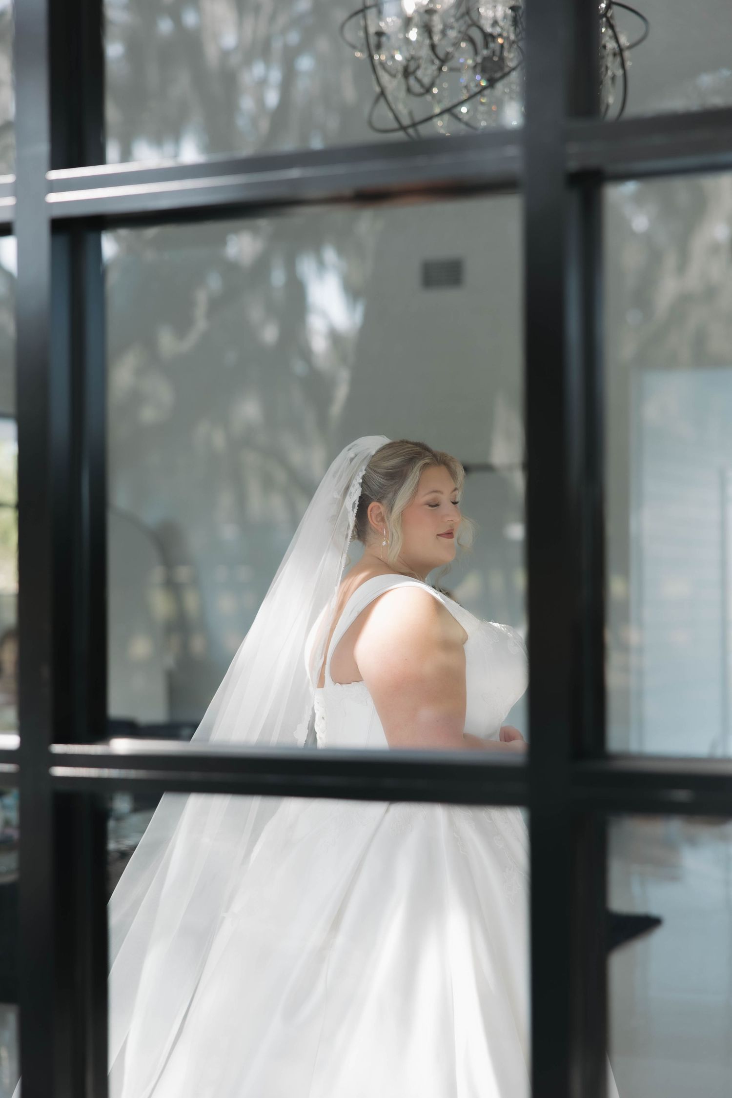 A bride in a white wedding dress and veil leans against a glass window in an architectural setting.