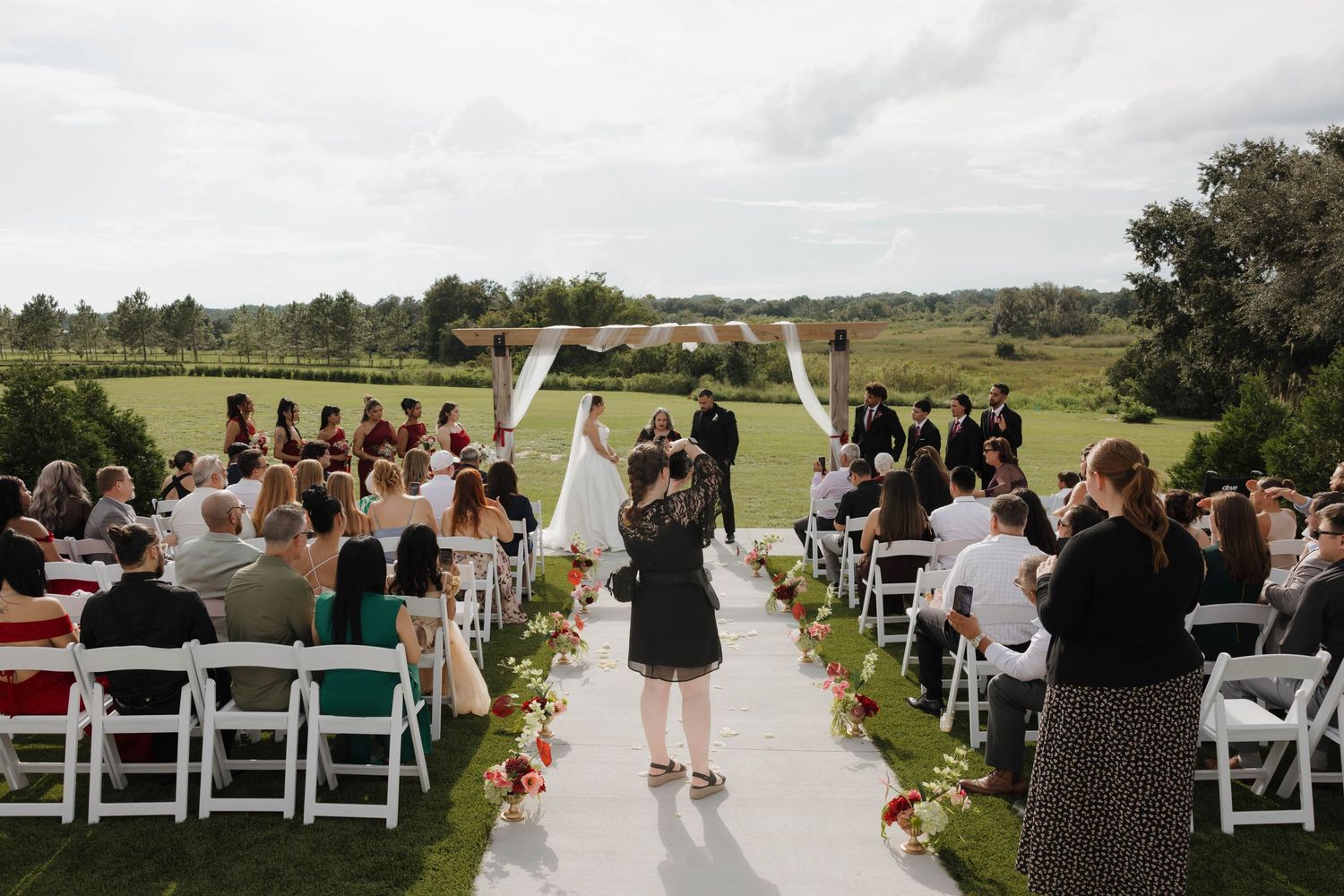 Outdoor wedding ceremony on lush green golf course with white chairs, floral aisle, arch, and guests gathered under cloudy sky.