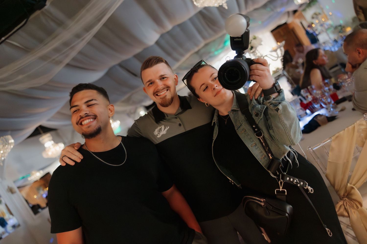 Three young men smiling at a wedding reception, one holding a professional video camera in a decorated venue.