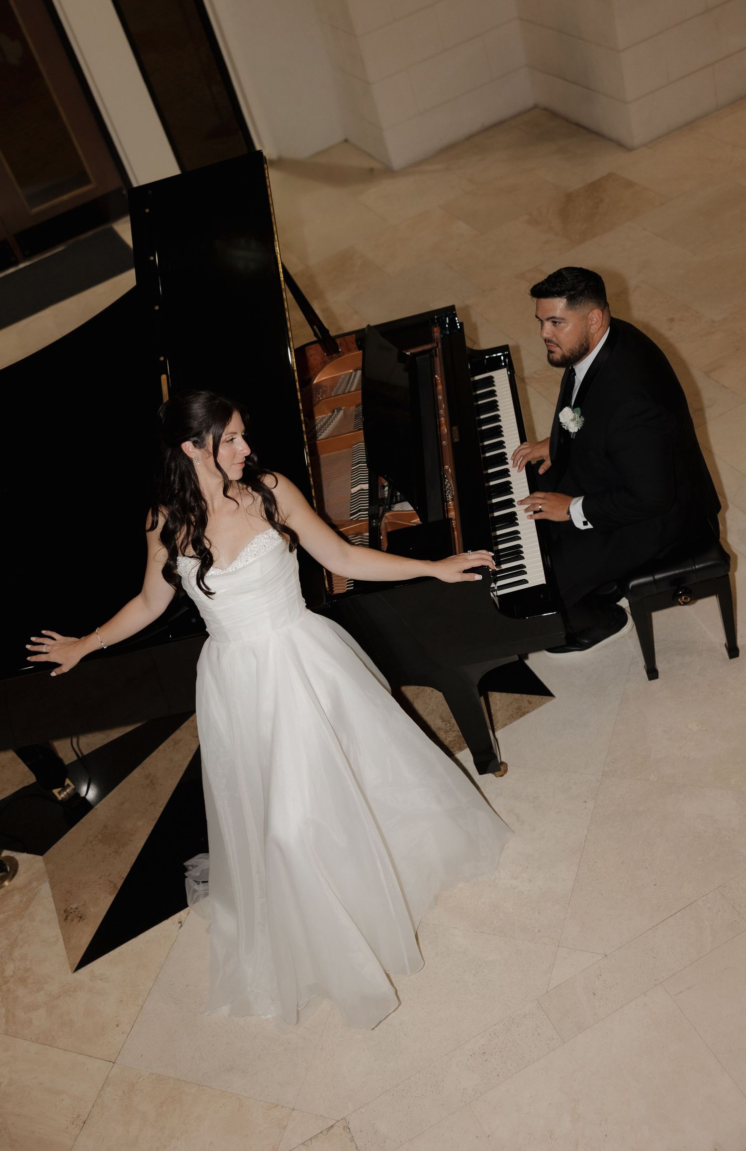 A bride in a white wedding gown dances playfully near a grand piano during a wedding reception.
