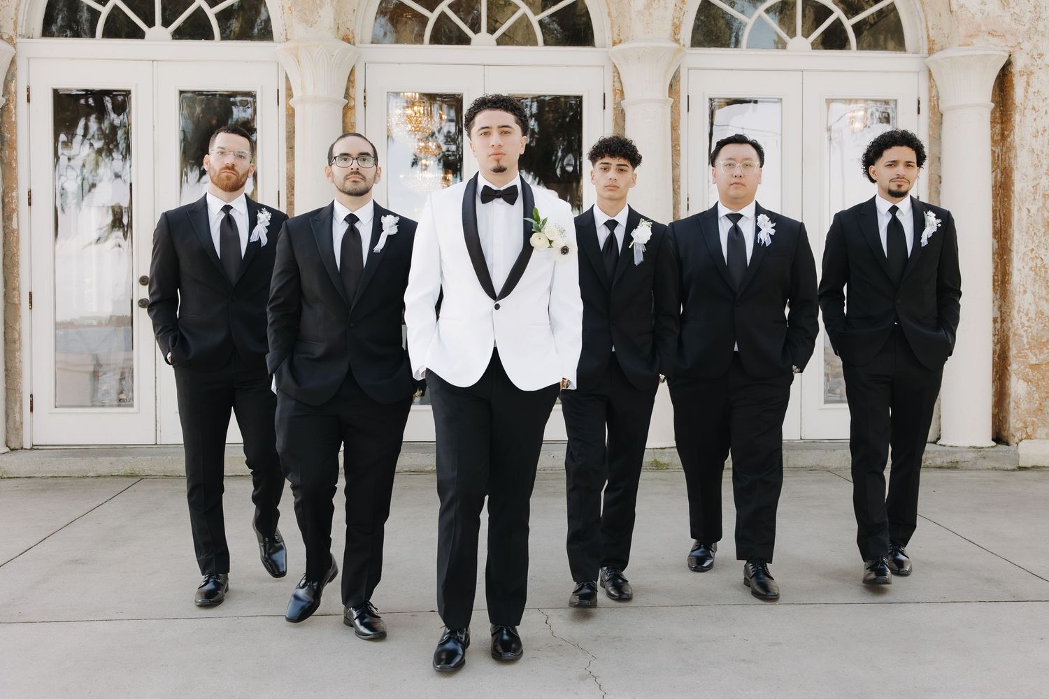 Groom in white tuxedo jacket stands with six groomsmen in black suits outside elegant venue.
