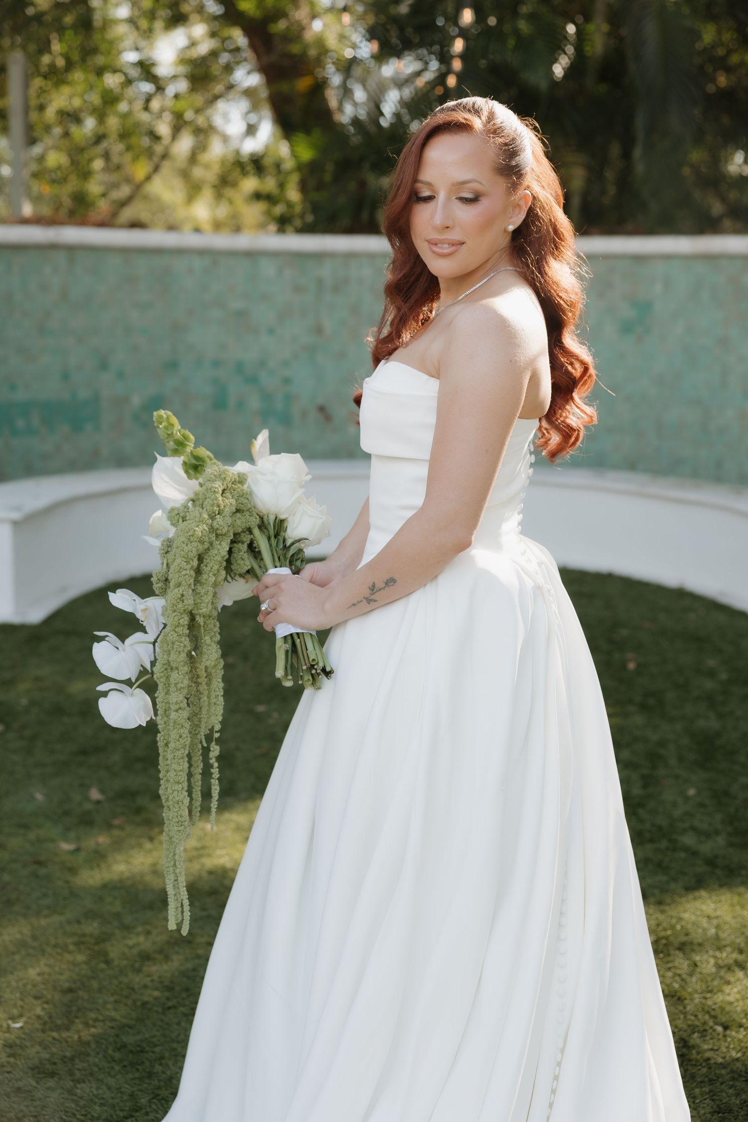 A bride in a flowing white wedding gown poses gracefully in an outdoor garden setting with greenery in the background.