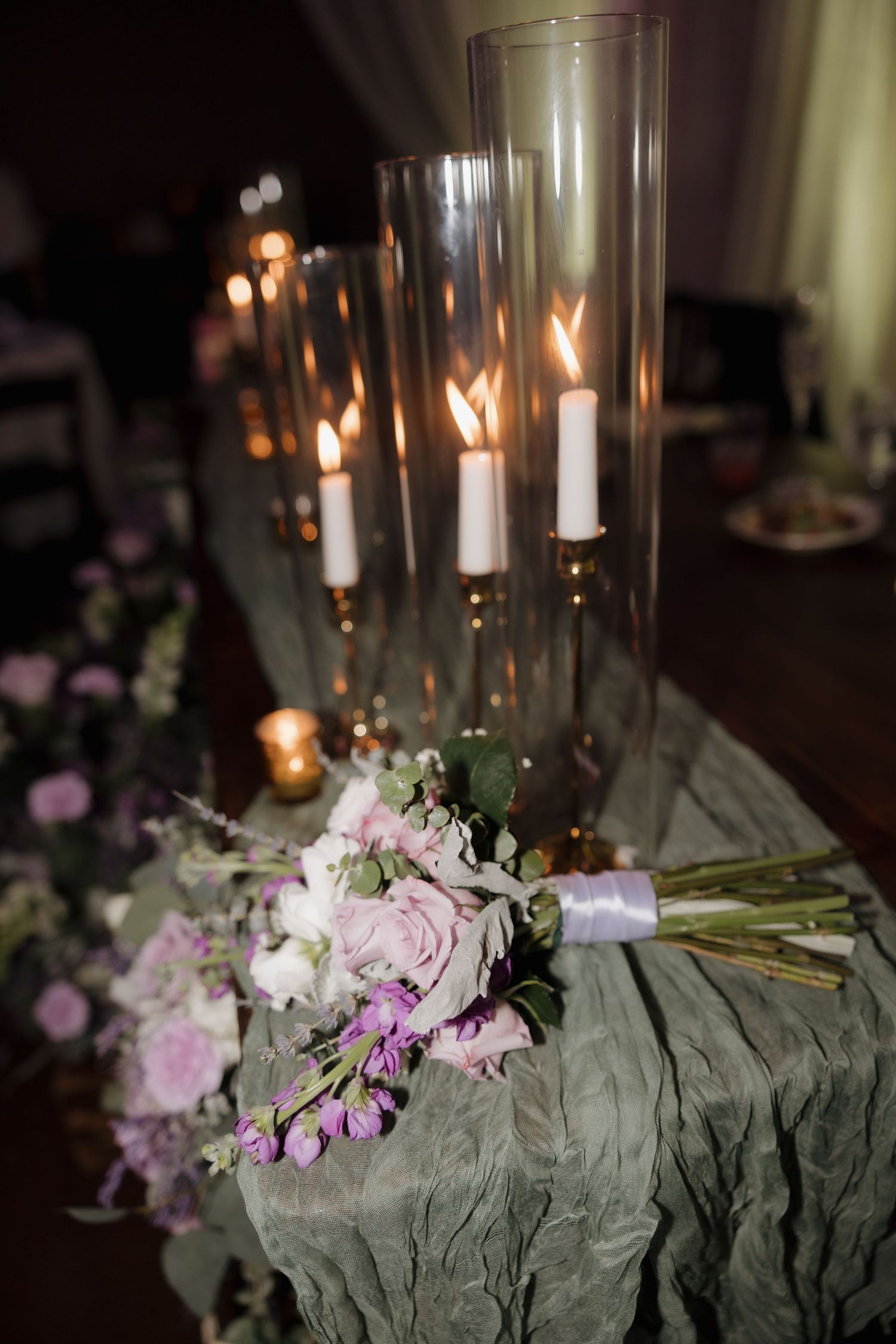 Elegant wedding table decor features lit candles in glass holders alongside pink and white floral arrangements on gray linens.