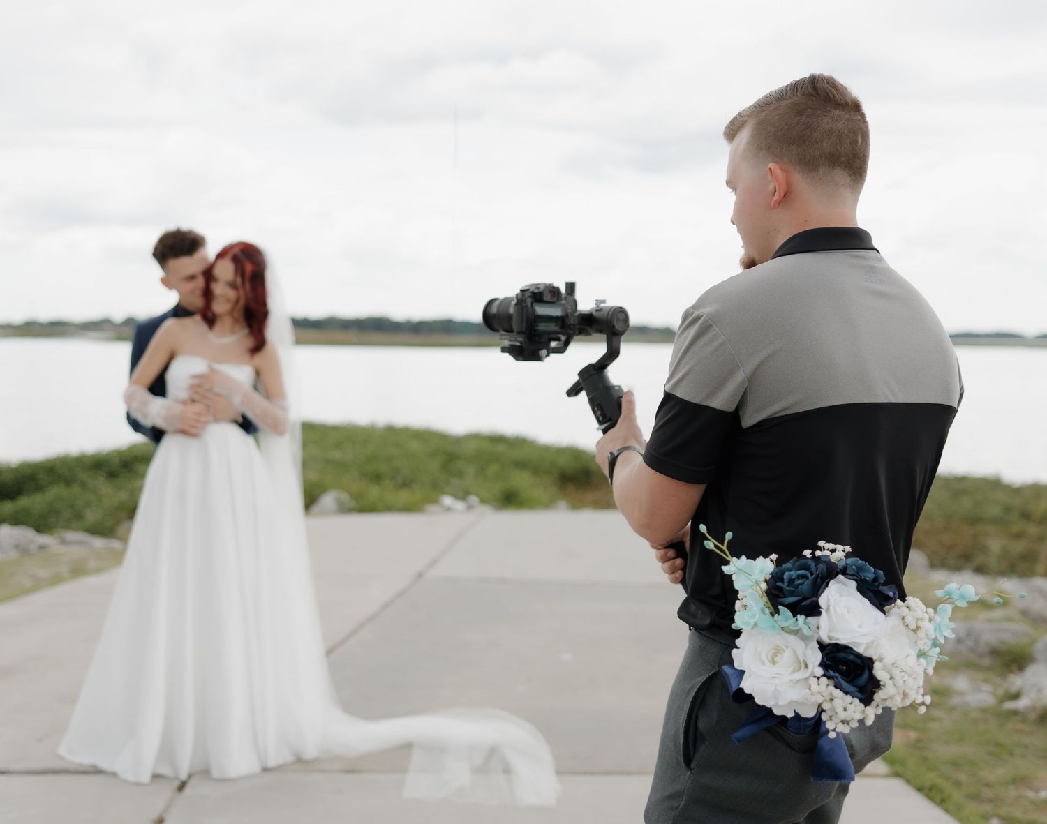 Videographer filming a bride and groom posing near a waterfront on their wedding day.
