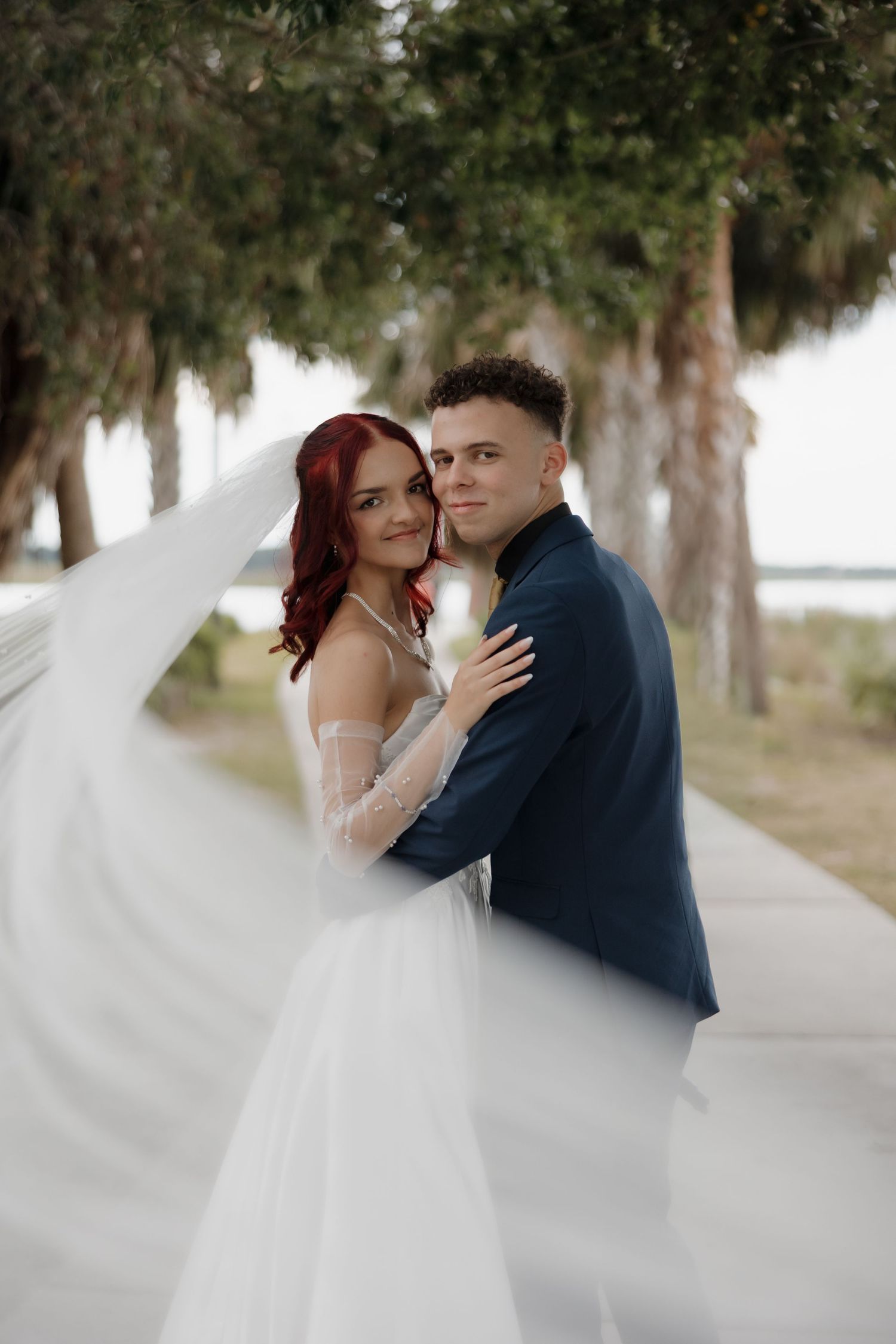 A bride in a flowing white gown and veil embraces her groom in a navy suit outdoors among tall trees.