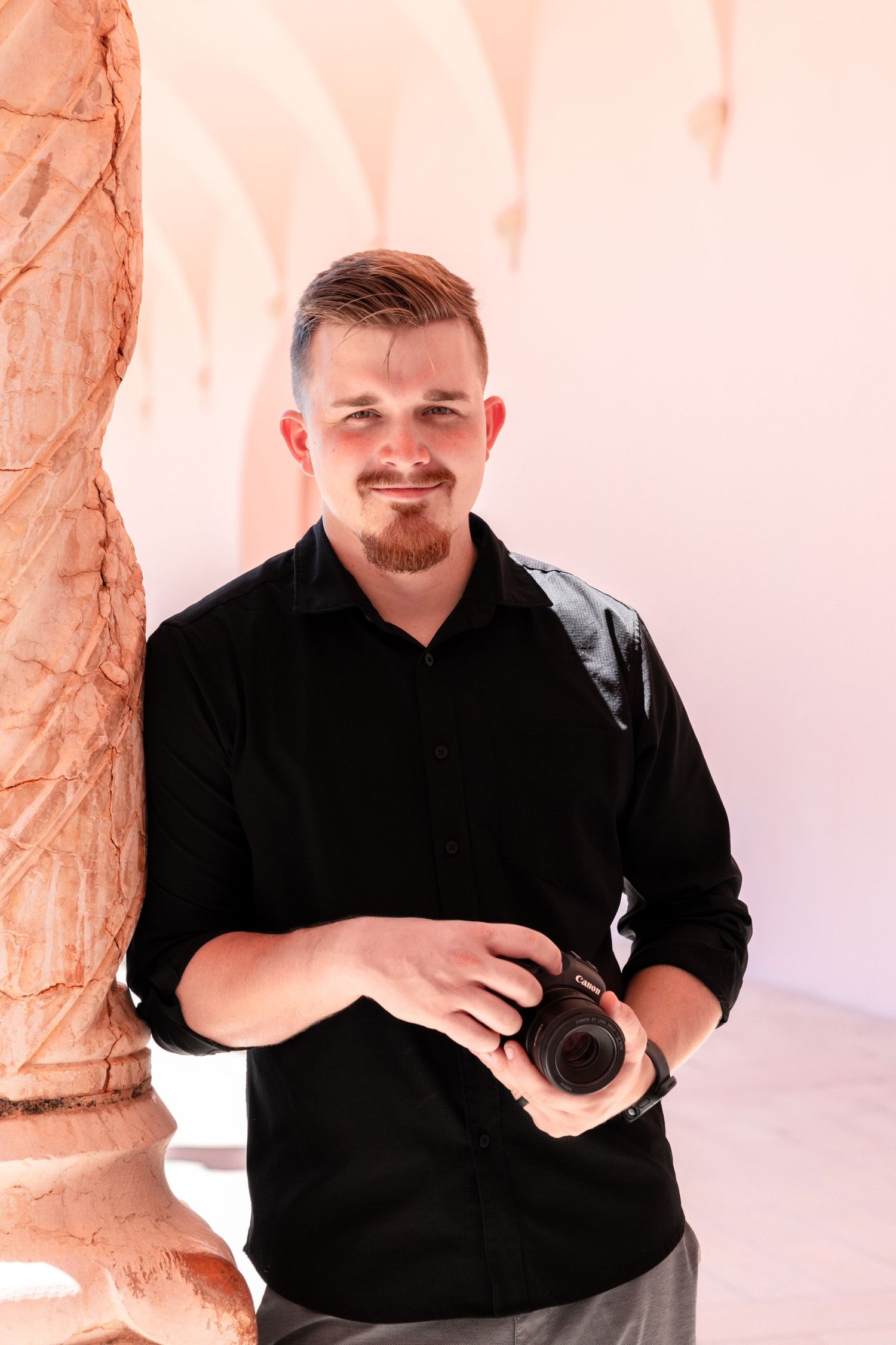 Young man with beard in black shirt holding DSLR camera, standing near ornate pink stone column.