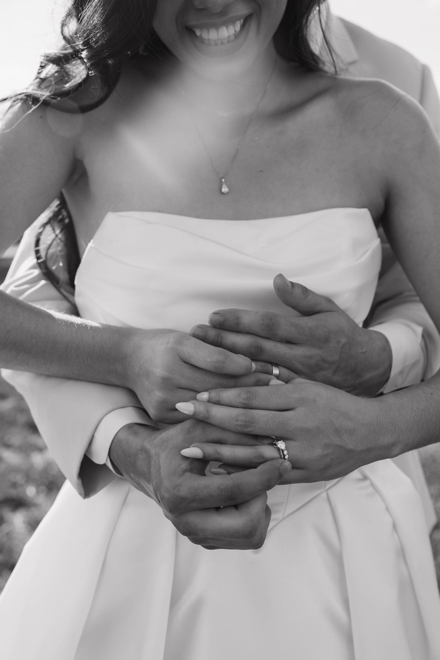 Black and white photo of bride and groom holding hands, showing wedding rings, bride in strapless gown smiling.