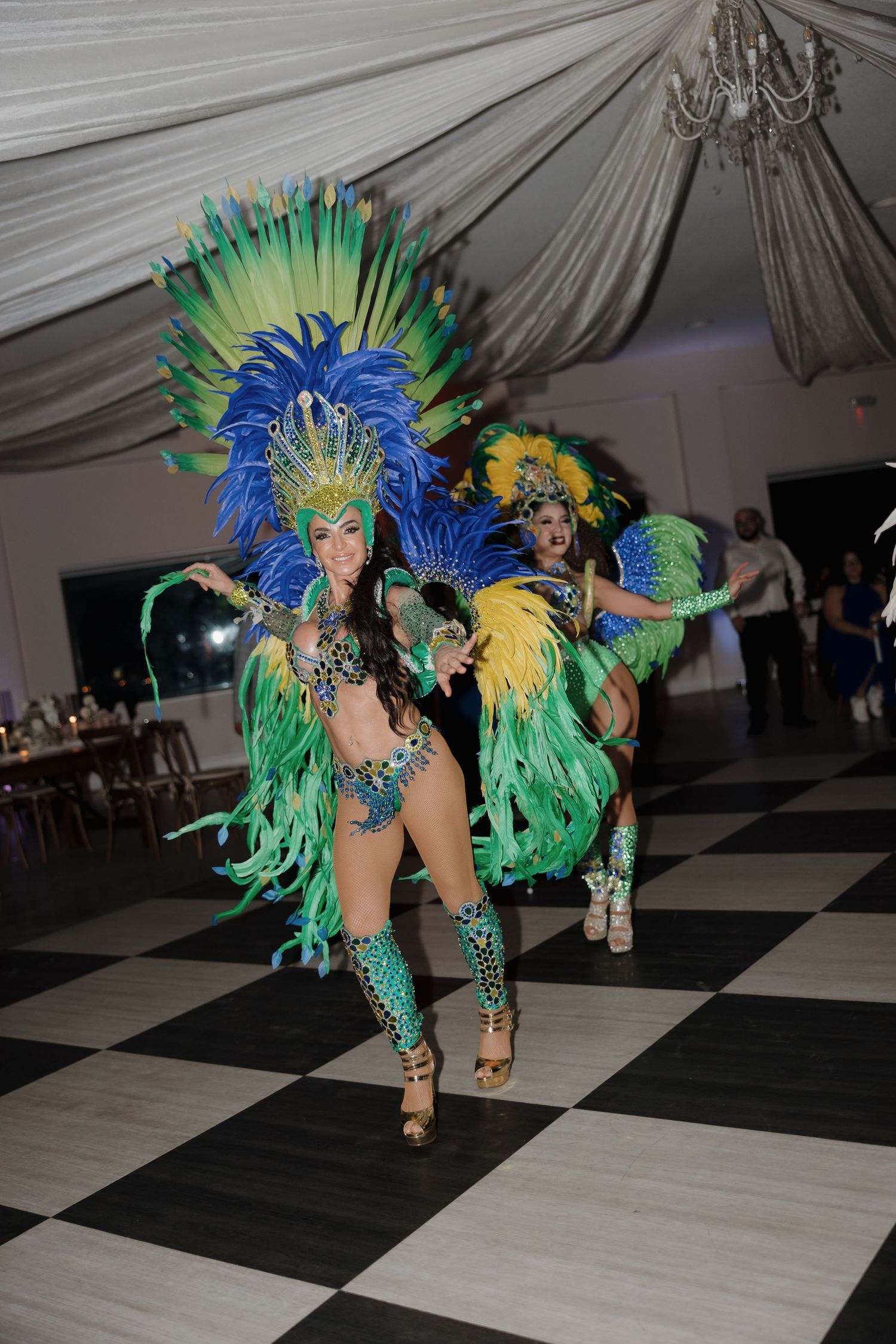 Carnival dancers in vibrant blue, green and yellow feathered costumes perform on a black and white checkered floor.