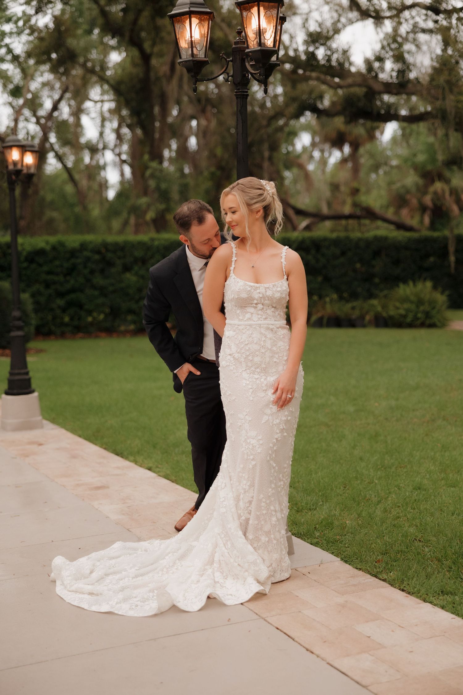 A couple shares a romantic moment on a garden path with vintage lantern posts and lush greenery in the background.