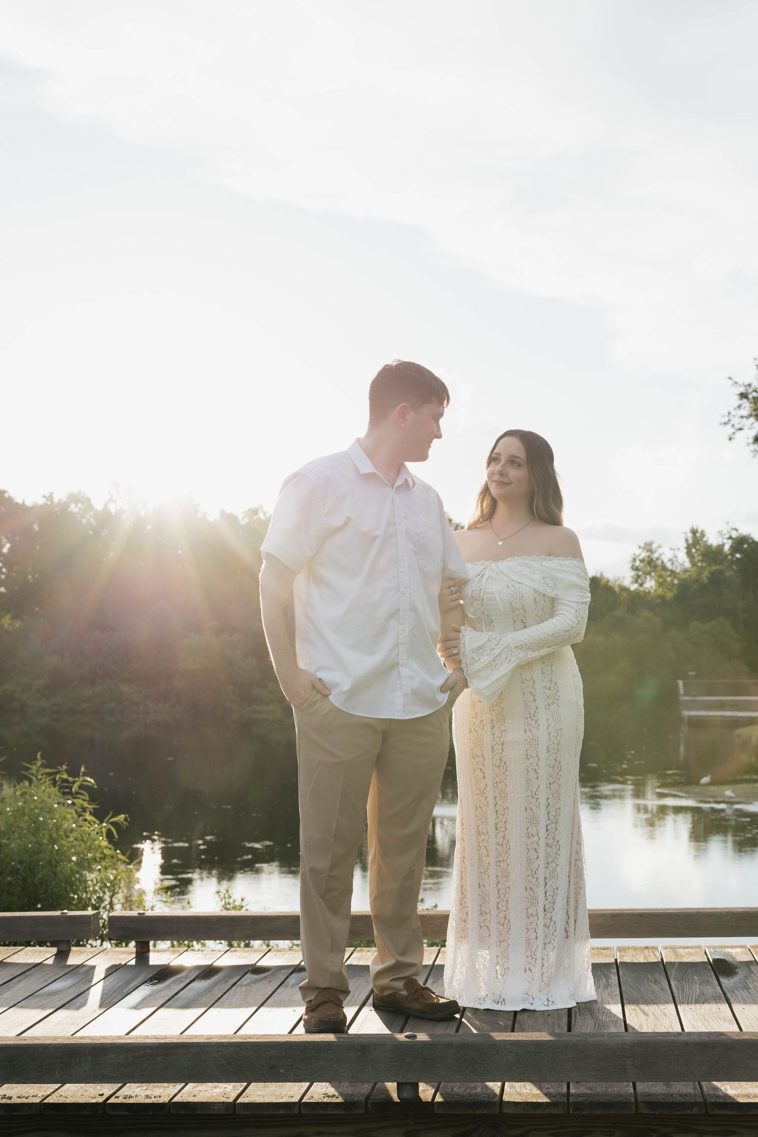 Couple standing on a wooden dock at golden hour, woman in white lace gown, man in white shirt, lake backdrop.