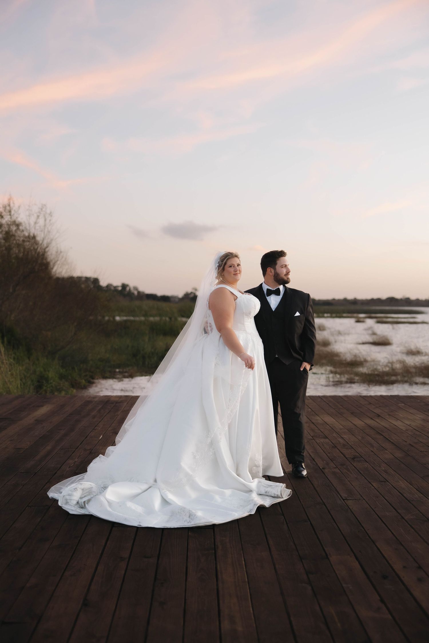 A couple in wedding attire poses together on a wooden boardwalk at sunset near the water.