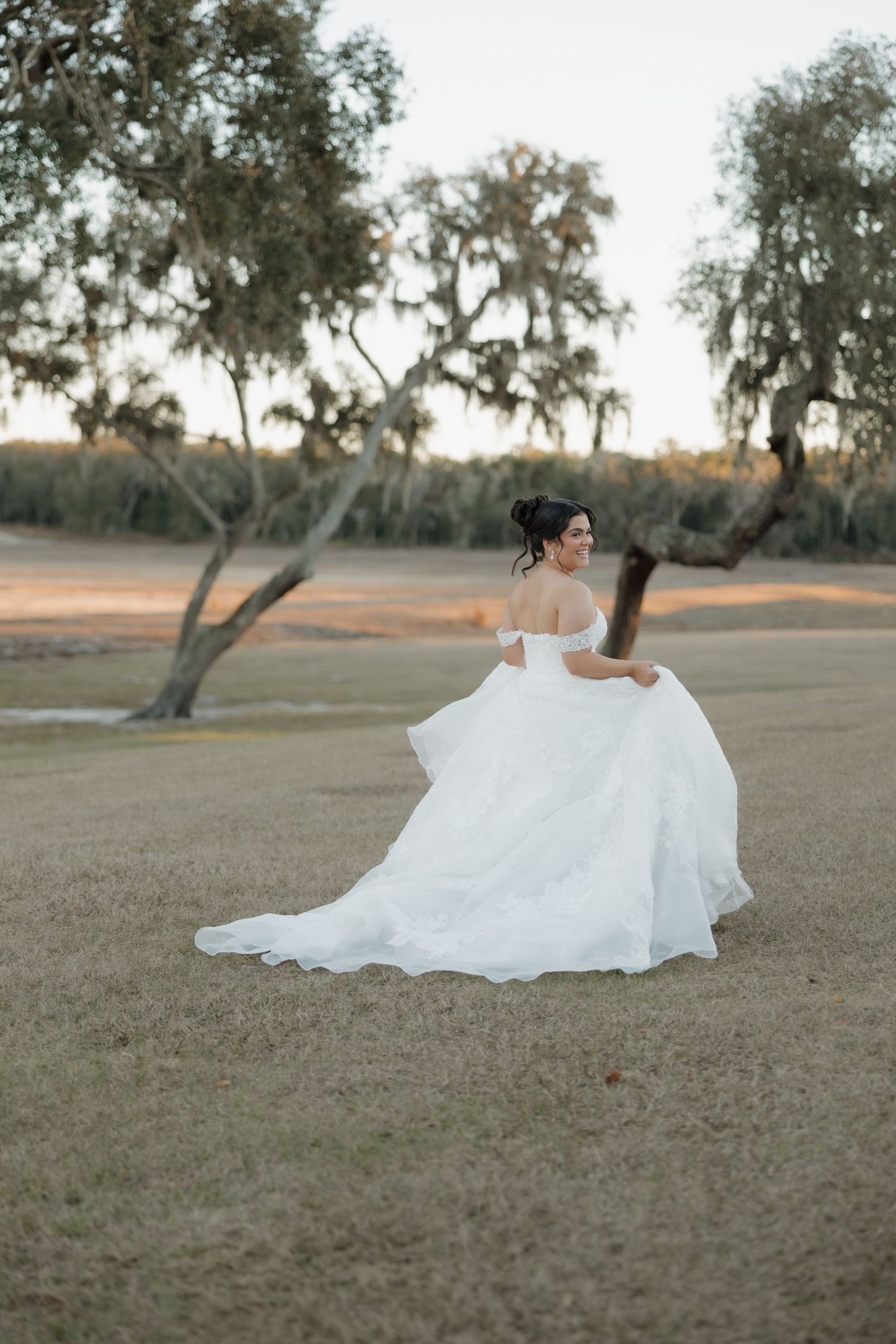 A flowing white wedding dress billows gracefully across a rustic outdoor field with olive trees in the background.
