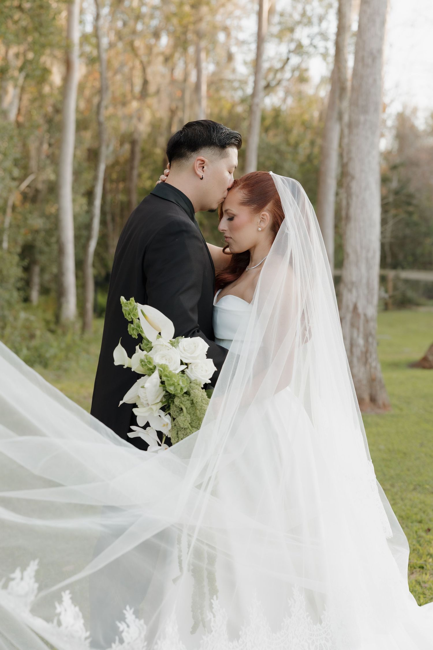 A romantic wedding couple shares a kiss outdoors surrounded by trees while the bride's veil flows in the breeze.
