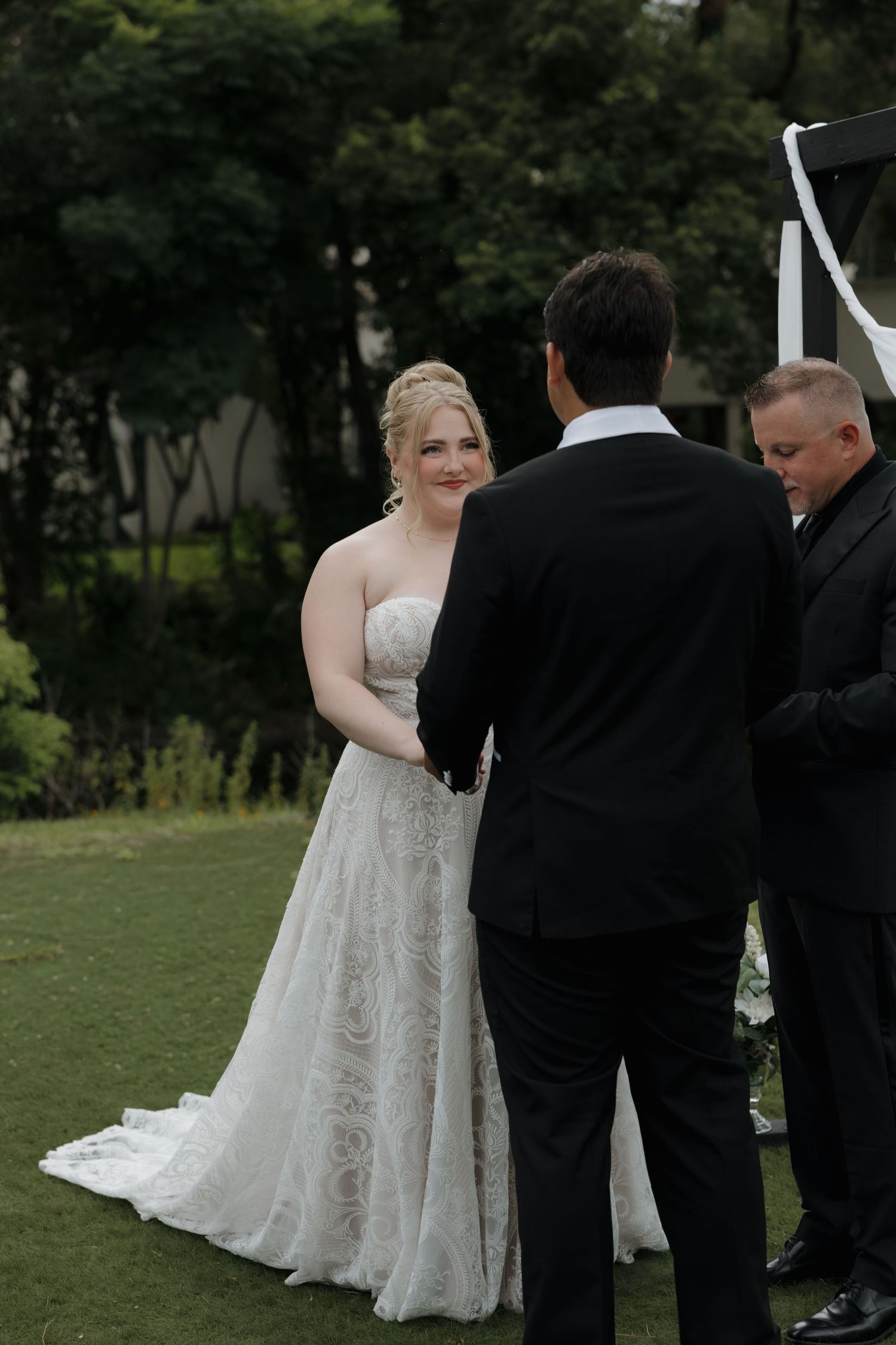Bride in lace gown smiling at groom in black tuxedo during outdoor wedding ceremony with officiant.