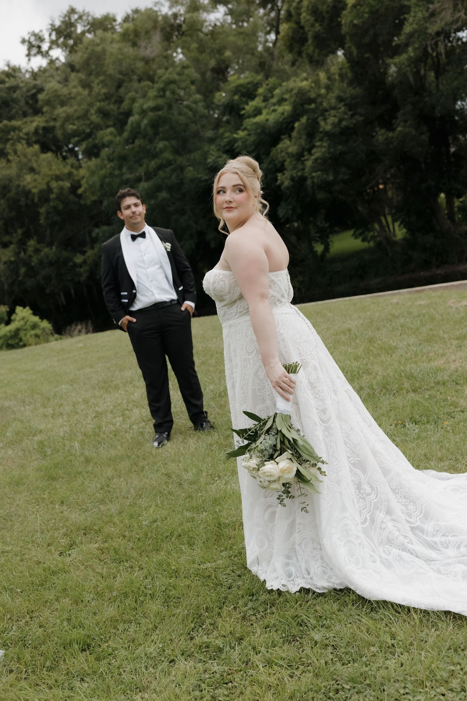 Bride in lace gown holding white bouquet with groom in tuxedo on green lawn at outdoor wedding.