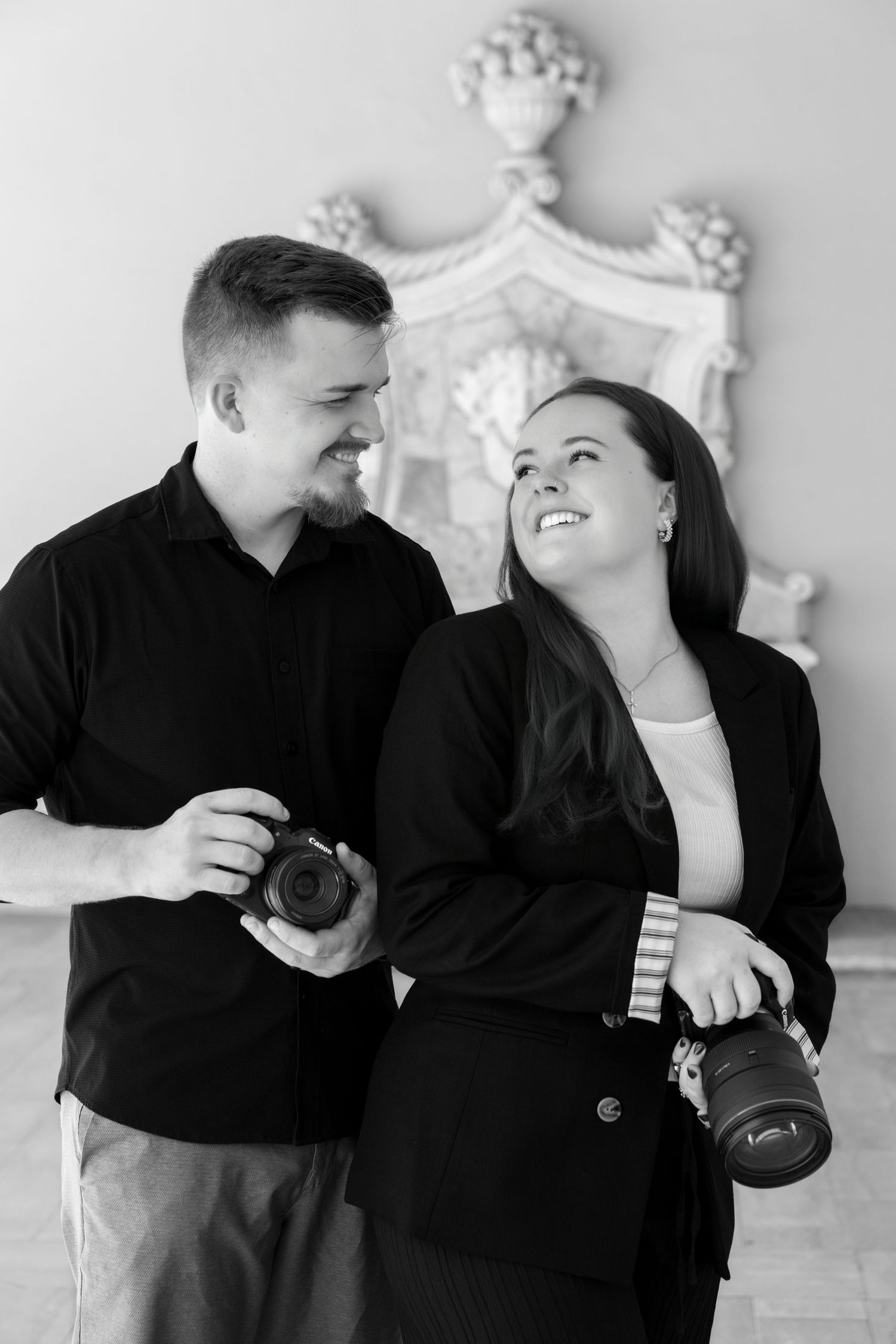 Black and white photo of a smiling couple holding cameras, standing together in an elegant room.