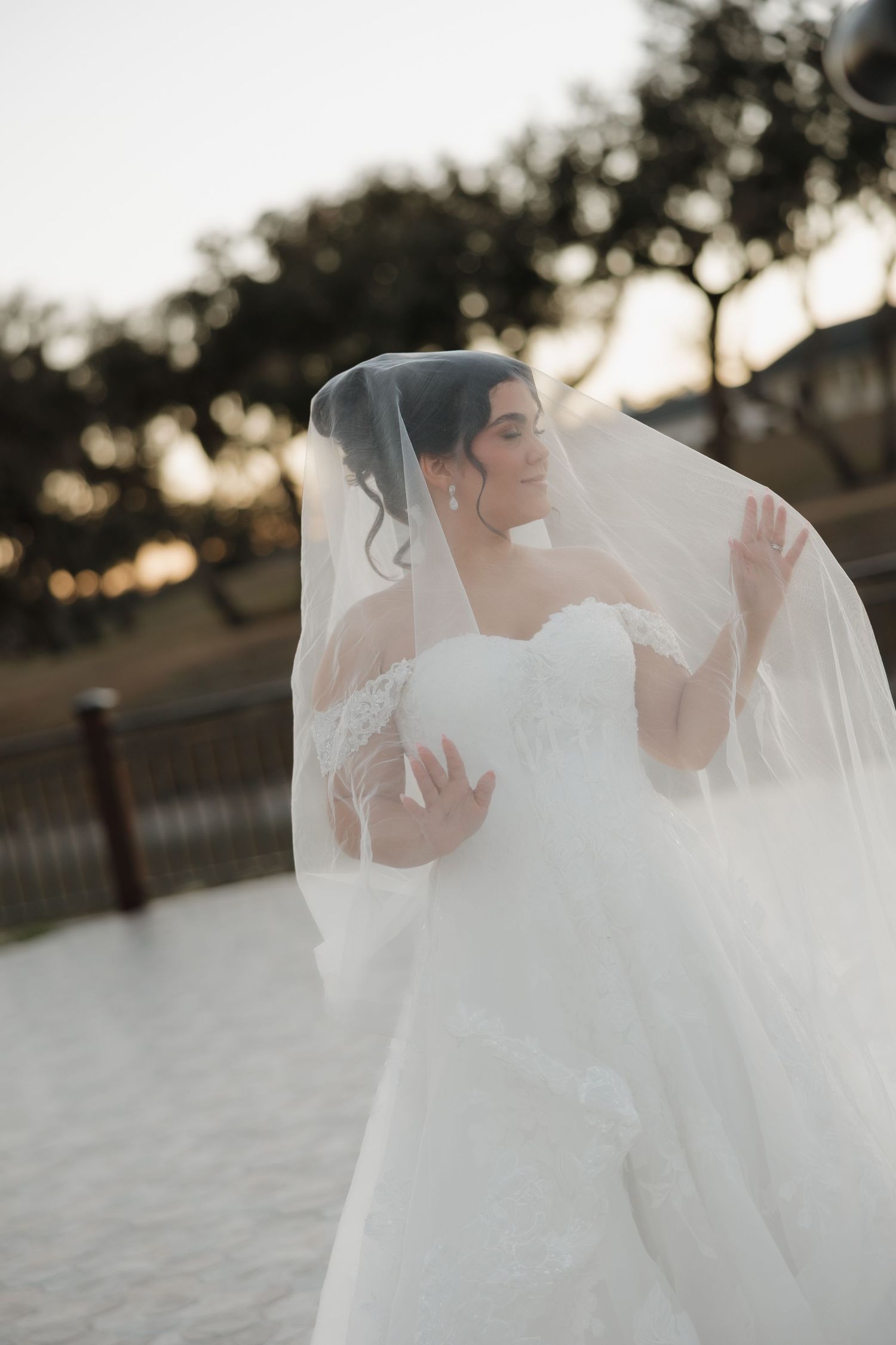 A bride in a flowing white wedding dress and veil stands gracefully in soft sunset lighting during an outdoor photoshoot.